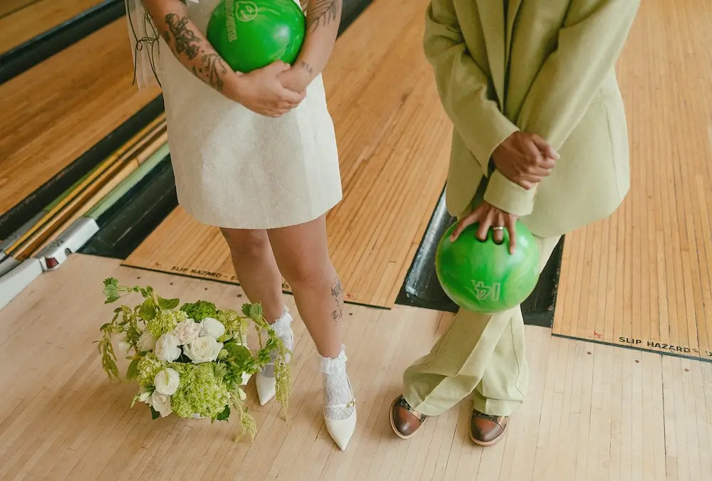 Detail of a couple holding green bowling balls on a vintage bowling lane, with a modern white wedding dress, sage suit, and green-and-white bouquet placed on the floor.