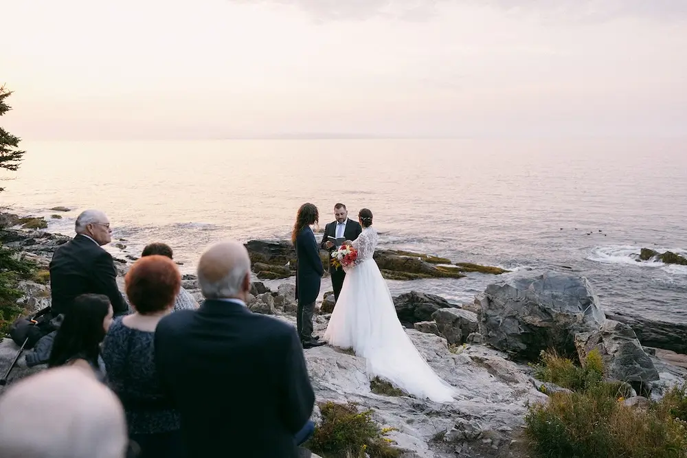 Small sunrise wedding ceremony on the rocky coastline of Acadia National Park with guests gathered nearby as the couple exchanges vows overlooking the ocean.