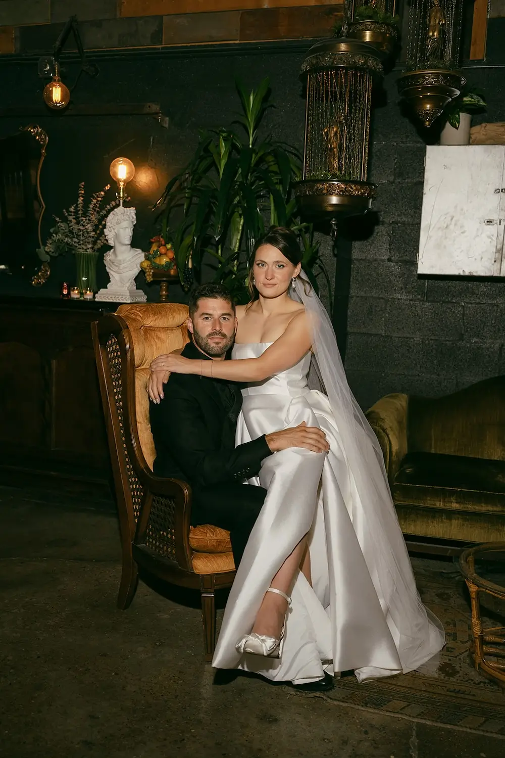 Bride in satin wedding gown sitting on groom’s lap in vintage lounge with plants, candles, and antique decor.