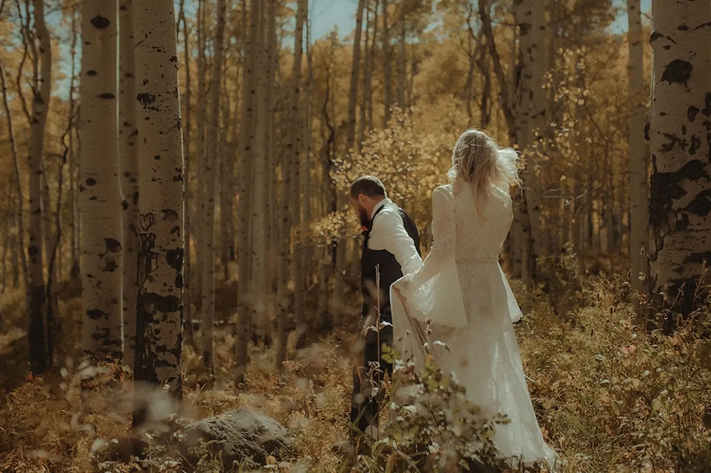 Bride and groom walking through golden aspen forest during autumn wedding.