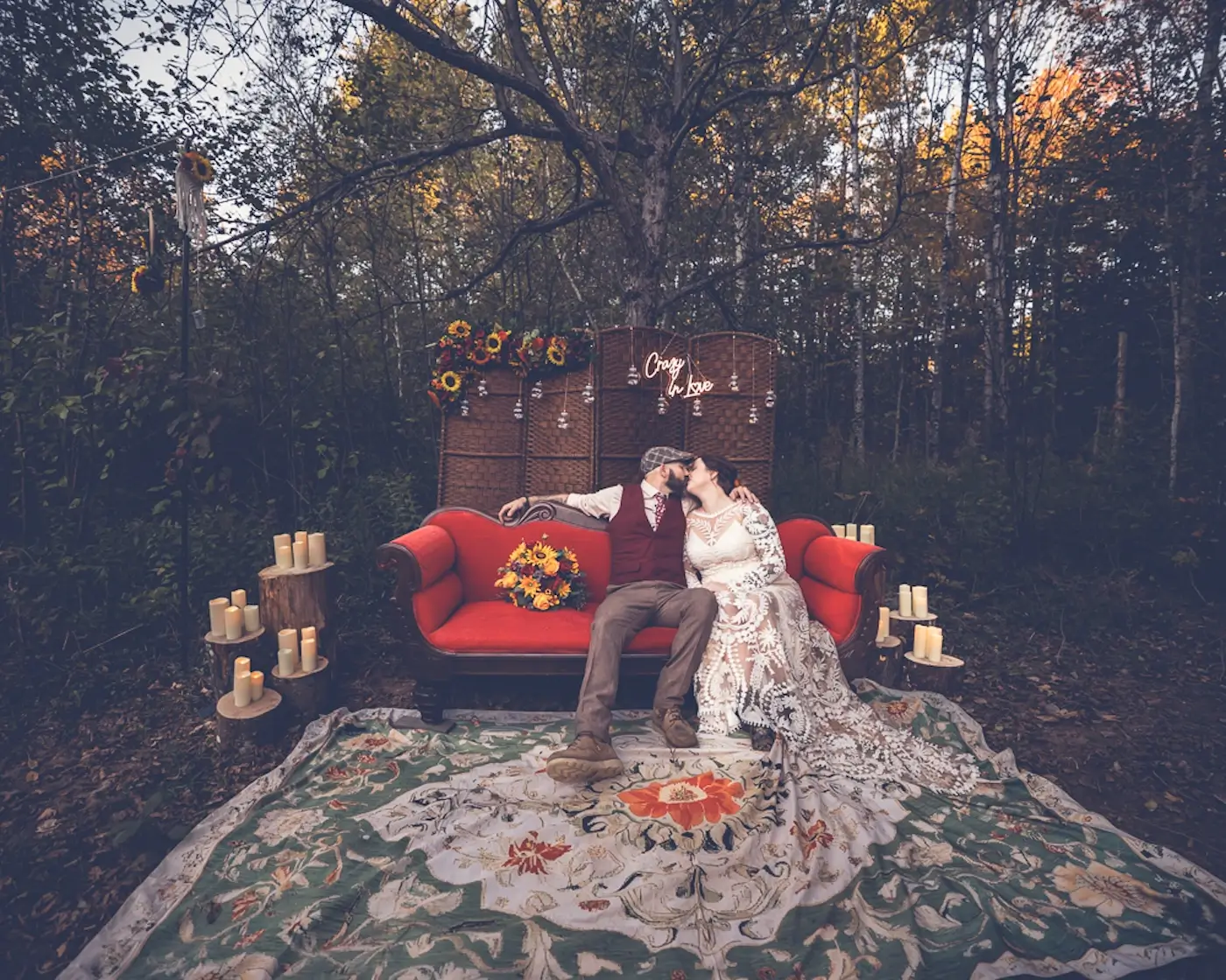 Bride and groom kissing on red velvet sofa atop vintage patterned rug with floral backdrop in forest wedding lounge.