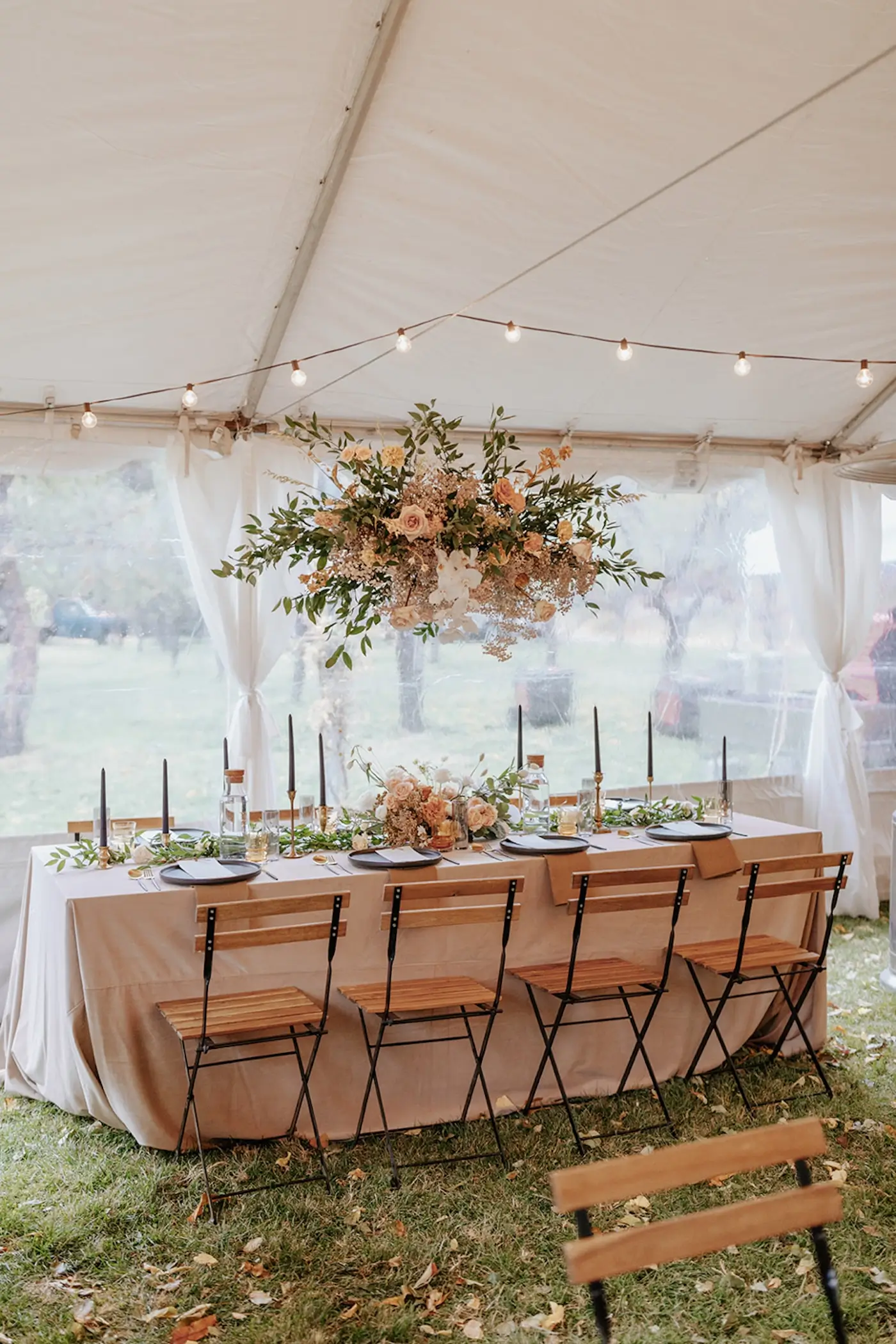 Boho wedding reception table under white tent with hanging floral installation, greenery, candles, and wooden chairs.