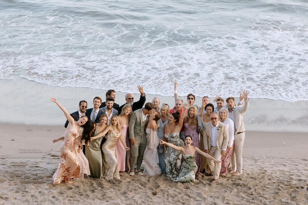 Beach micro wedding group portrait of a bride and groom embracing surrounded by family and friends in cocktail attire, ocean waves behind them