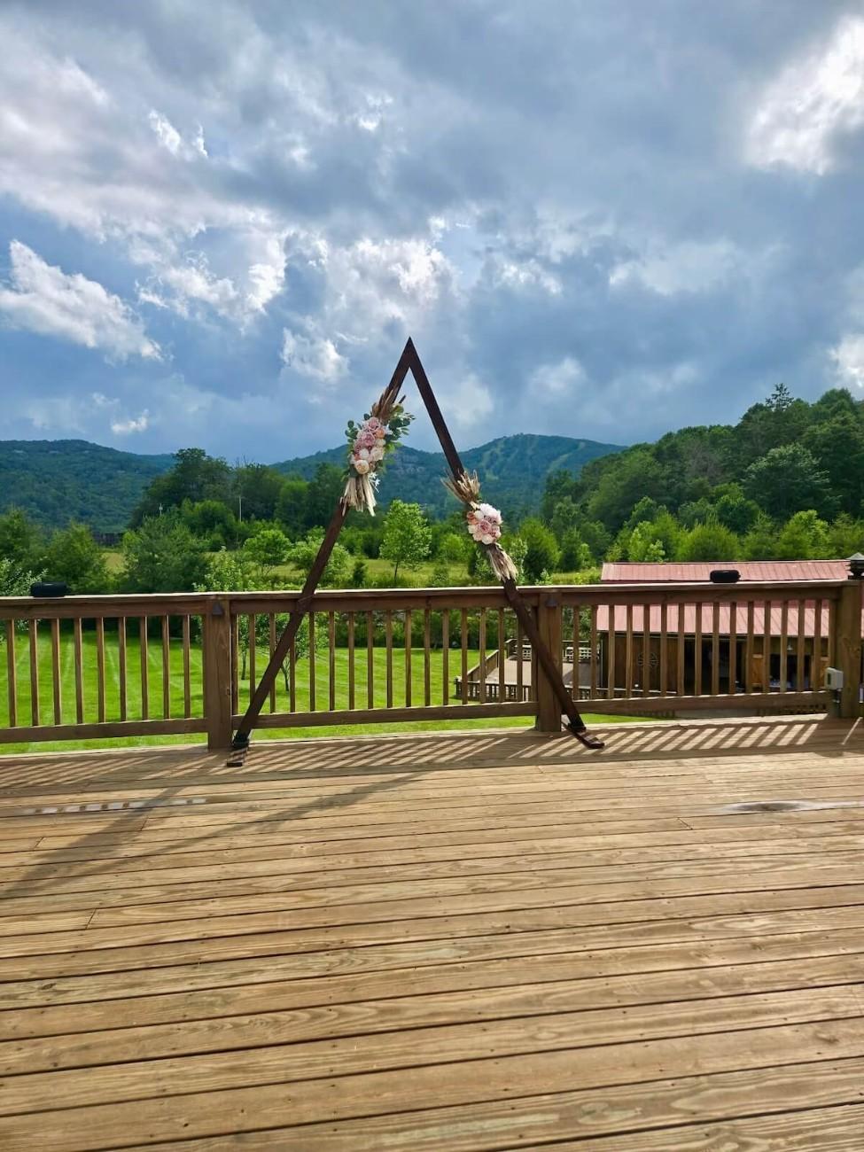 Triangular wood ceremony arch on a Banner Elk deck overlooking Blue Ridge pasture and forested ridges under a cloudy summer sky