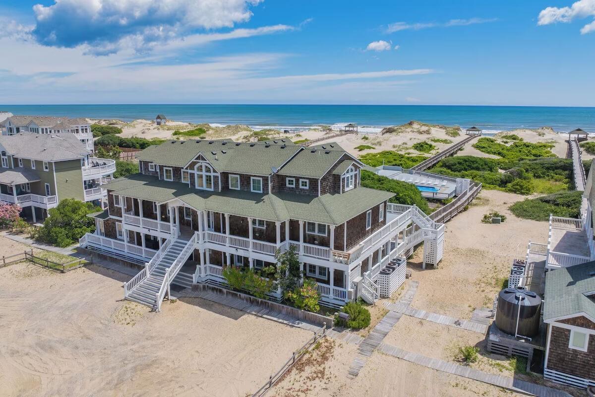 Aerial of a cedar-shake Outer Banks VRBO wedding venue with wraparound porches and private boardwalk across sea grass to the beach, Atlantic to the horizon