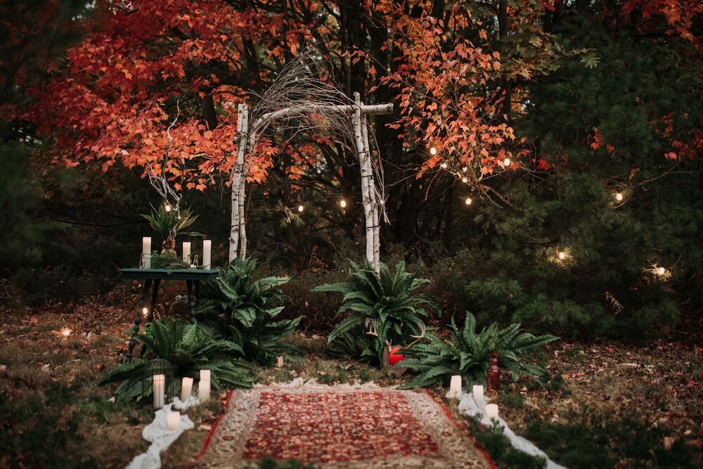 Twilight autumn ceremony — birch-log arch under fiery red maples, potted ferns flanking a red oriental aisle runner with trailing lace and lanterns