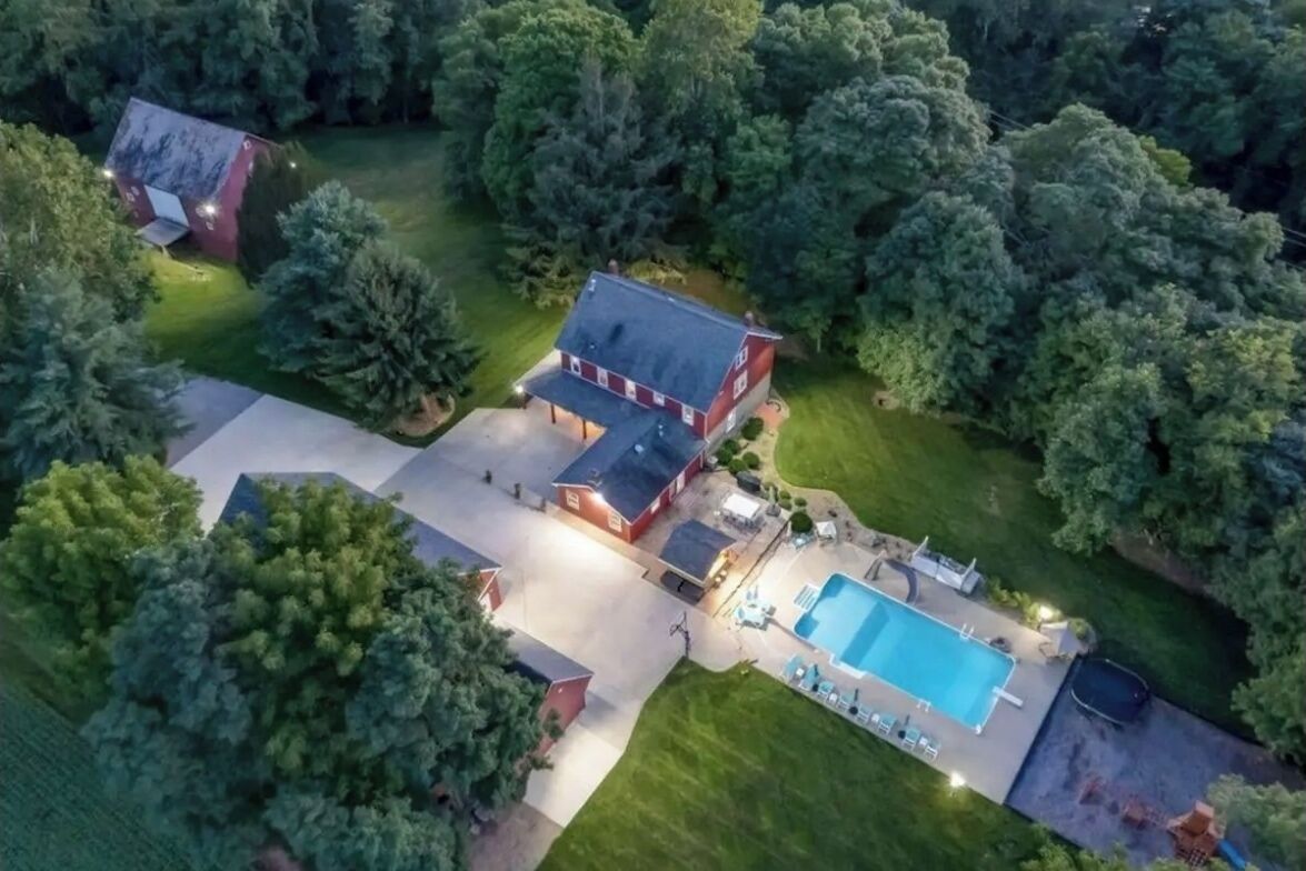 Aerial of a red gambrel-roof Ohio VRBO wedding venue at blue hour, rectangular pool and stone patio, mature shade trees framing the property