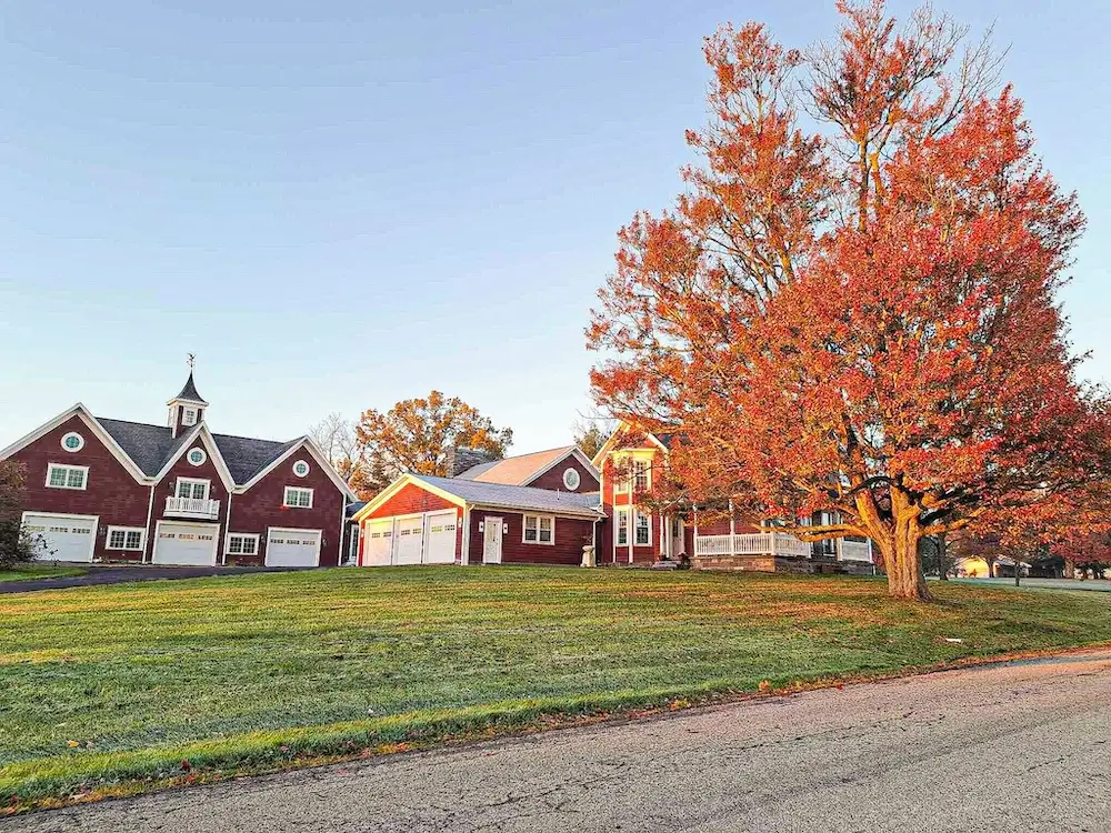 Red carriage house with white cupola and attached garages, red main house with pillared porch, autumn maple in red foliage between at golden hour