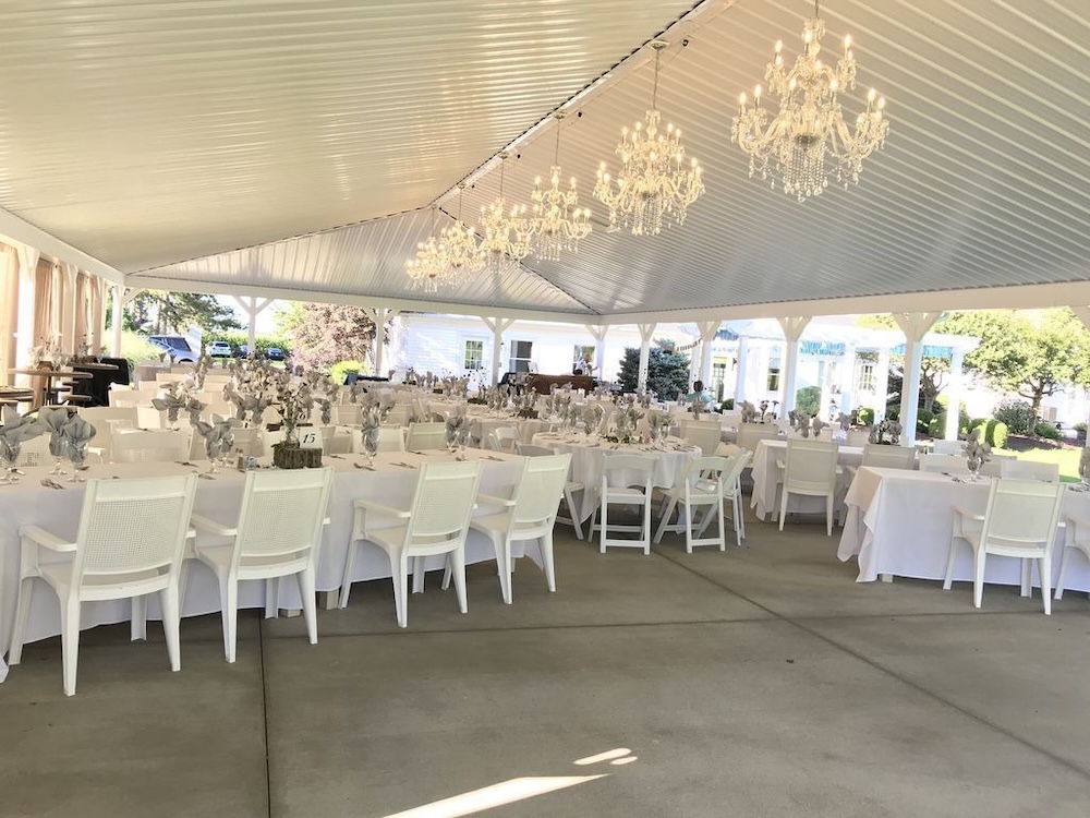 Open-air reception pavilion with white-painted ceiling and four crystal chandeliers, long white-linen tables set for a wedding dinner, lawn beyond