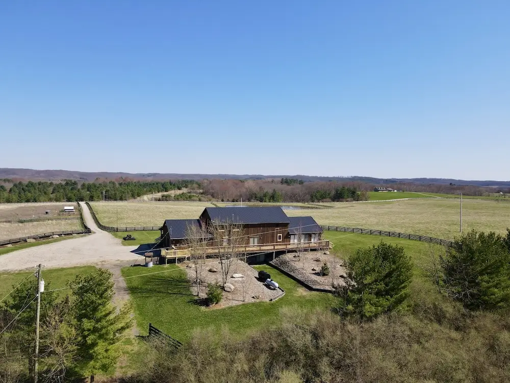 Dark-stained wood Hocking Hills lodge on a hilltop, vast Ohio pastureland and tree-covered ridges, dirt drive approaching, split-rail fence, open sky