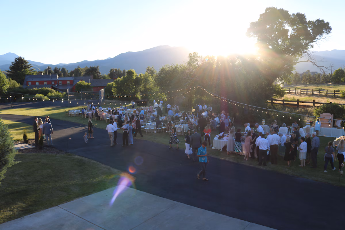 Outdoor reception at golden hour — guests at long tables under bistro string lights between mature trees, red barn at left, Wasatch silhouetted behind