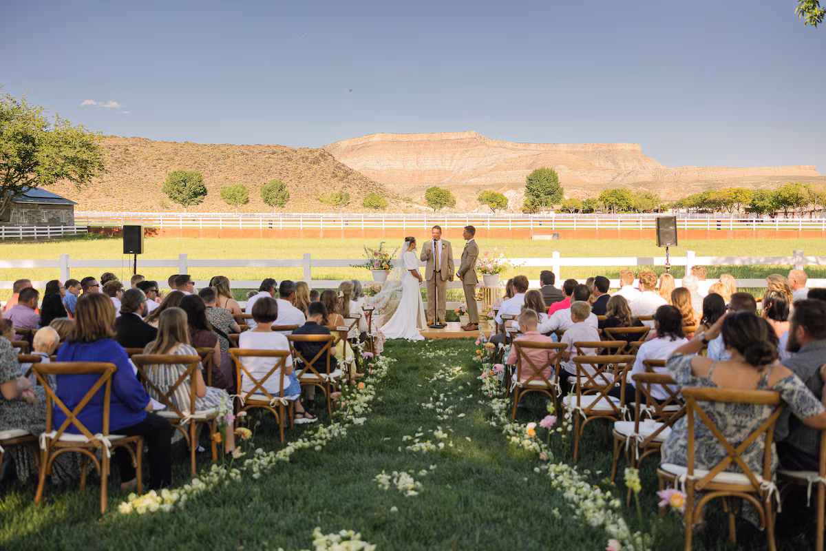 Outdoor ranch ceremony — couple at the altar, cross-back chairs flanking a petal-lined grass aisle, white horse fence, red rock mesa rising behind
