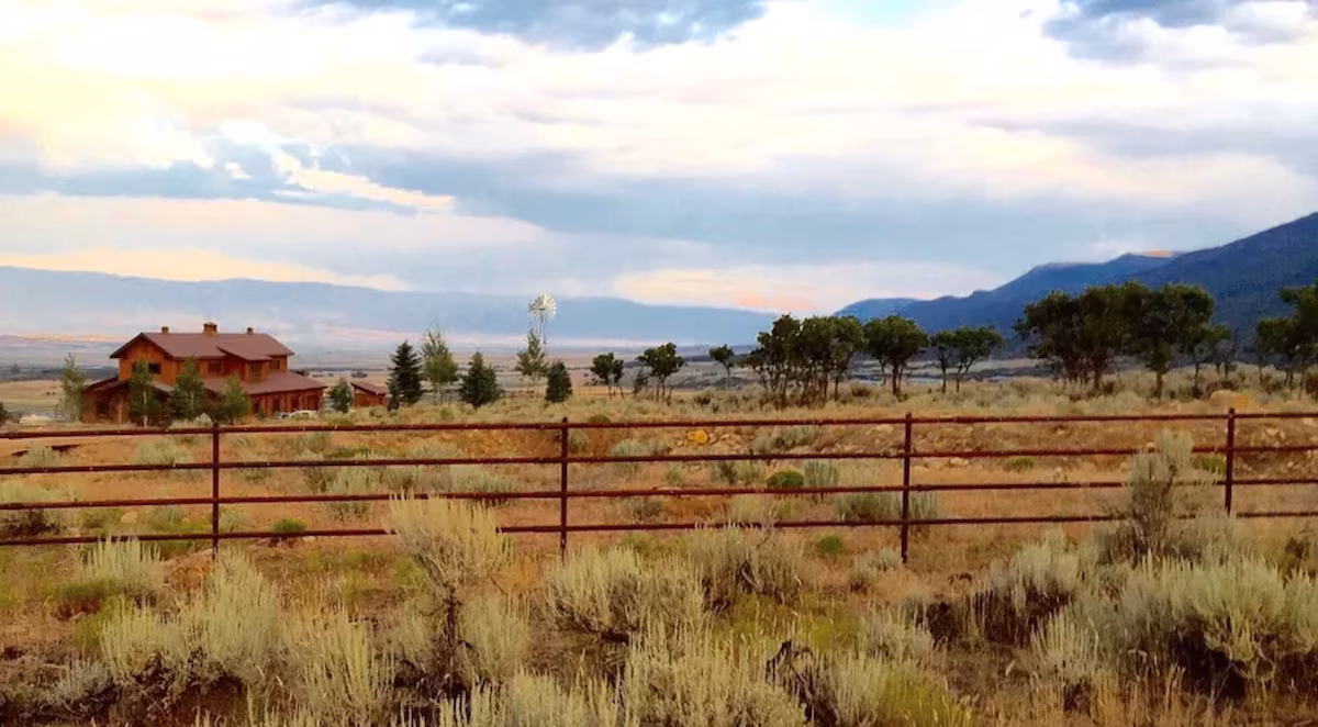Brown Utah VRBO wedding venue farmhouse on a sage prairie at sunset, distant windmill, mountains silhouetted under pink-blue sky, iron rail fence