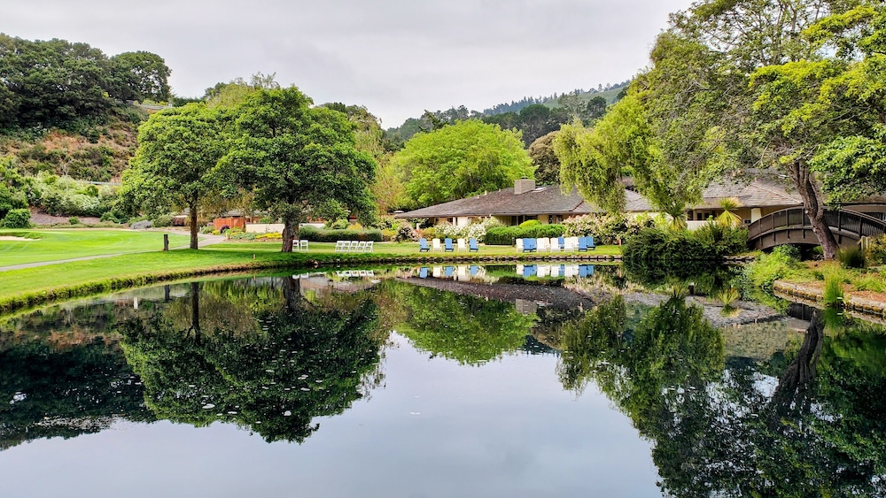 Mirror-still pond reflecting a Carmel Valley resort lodge, willows and oaks on the bank, blue Adirondack chairs on the lawn and a wooden footbridge
