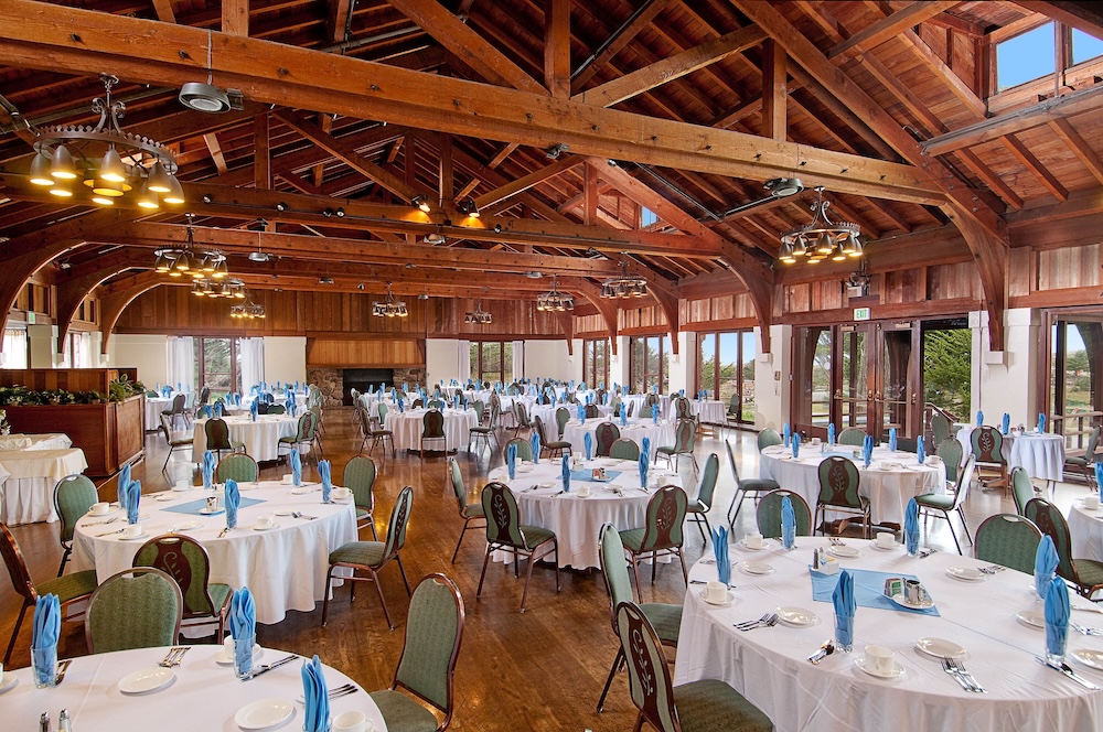 Cathedral-ceilinged lodge dining hall with exposed wood trusses, round tables in white linen and blue napkins, and a stone fireplace at the far end