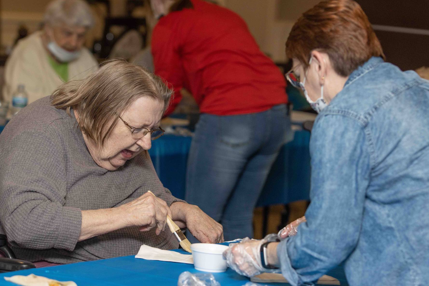 A senior woman using a basting brush to cover something on a plate on a blue table.