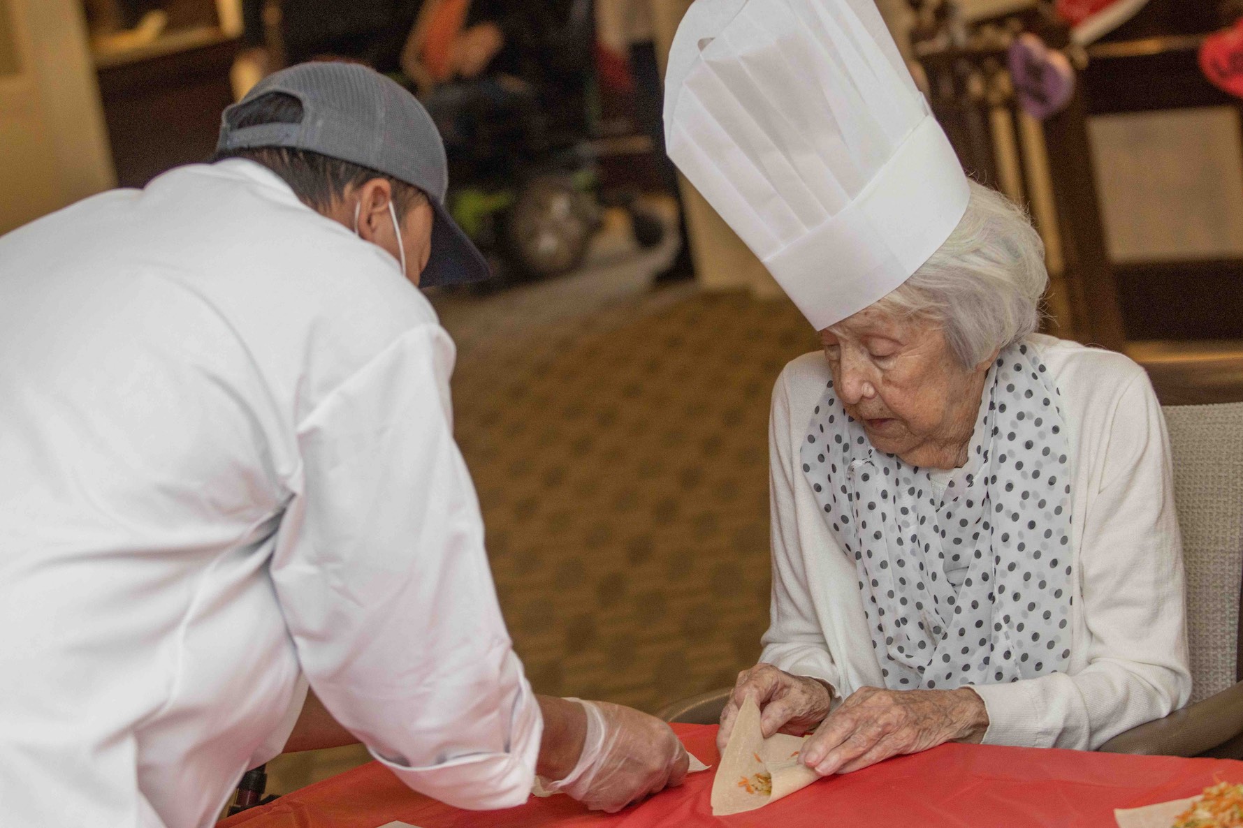 A senior woman in a chef's hat working with another chef to make lobster rolls on a red table.