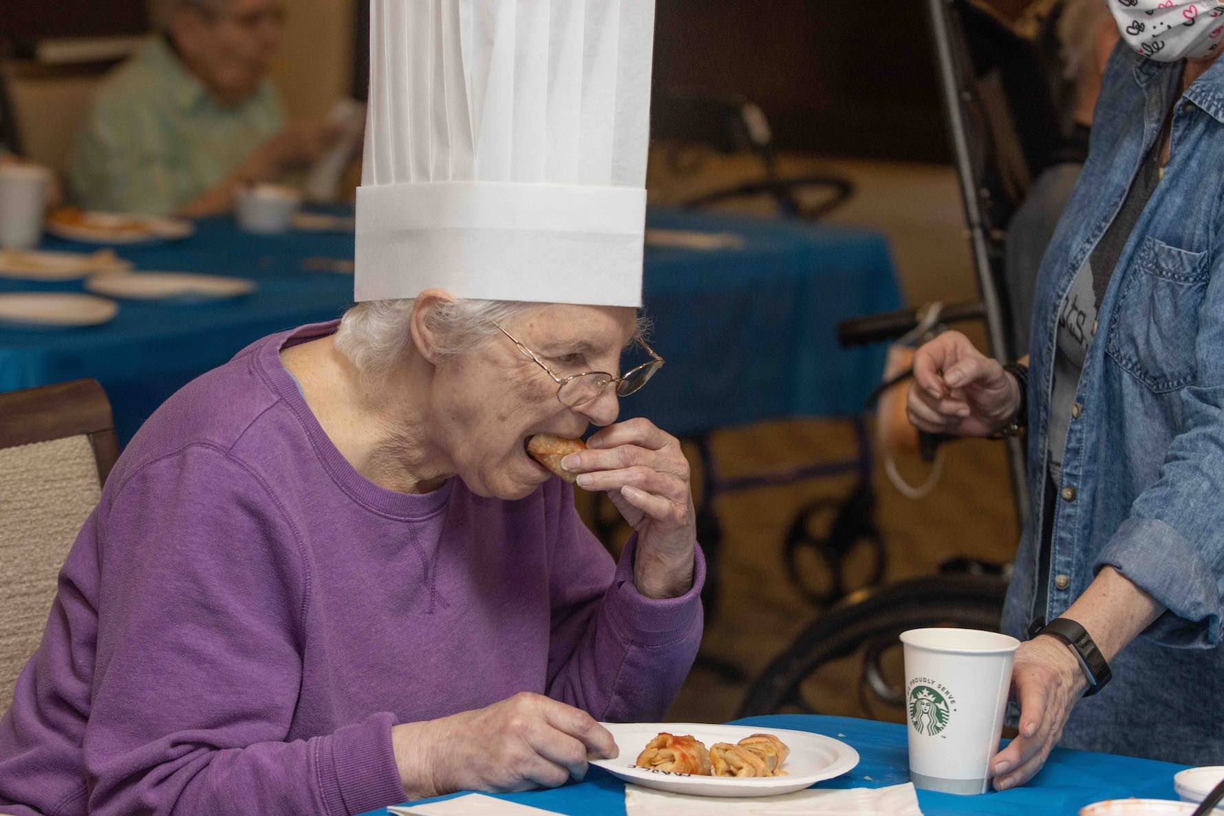 A woman in a white chef's hat and lavender long-sleeve shirt taking a bite of a pastry.