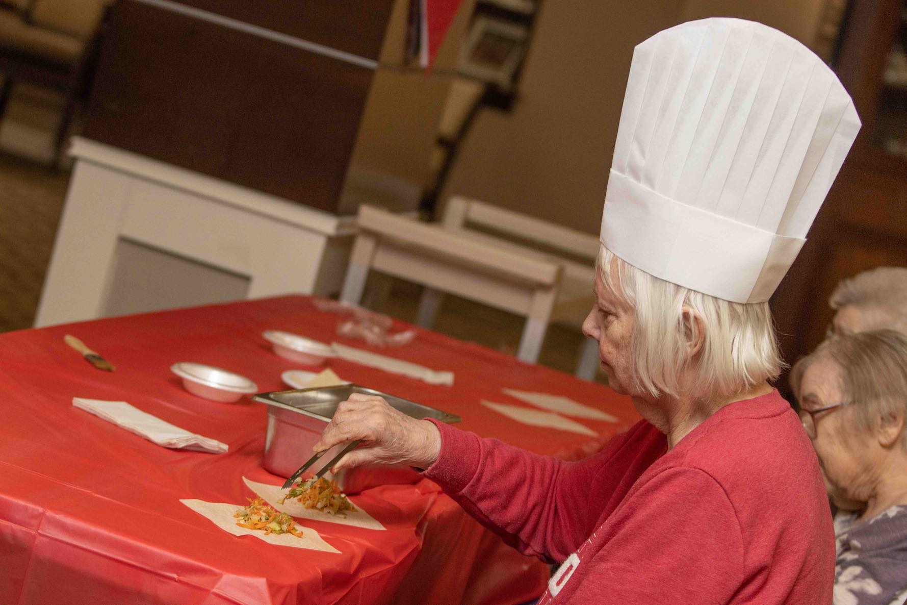 A woman in a white chef's hat using tongs to prepare food on a red tablecloth.