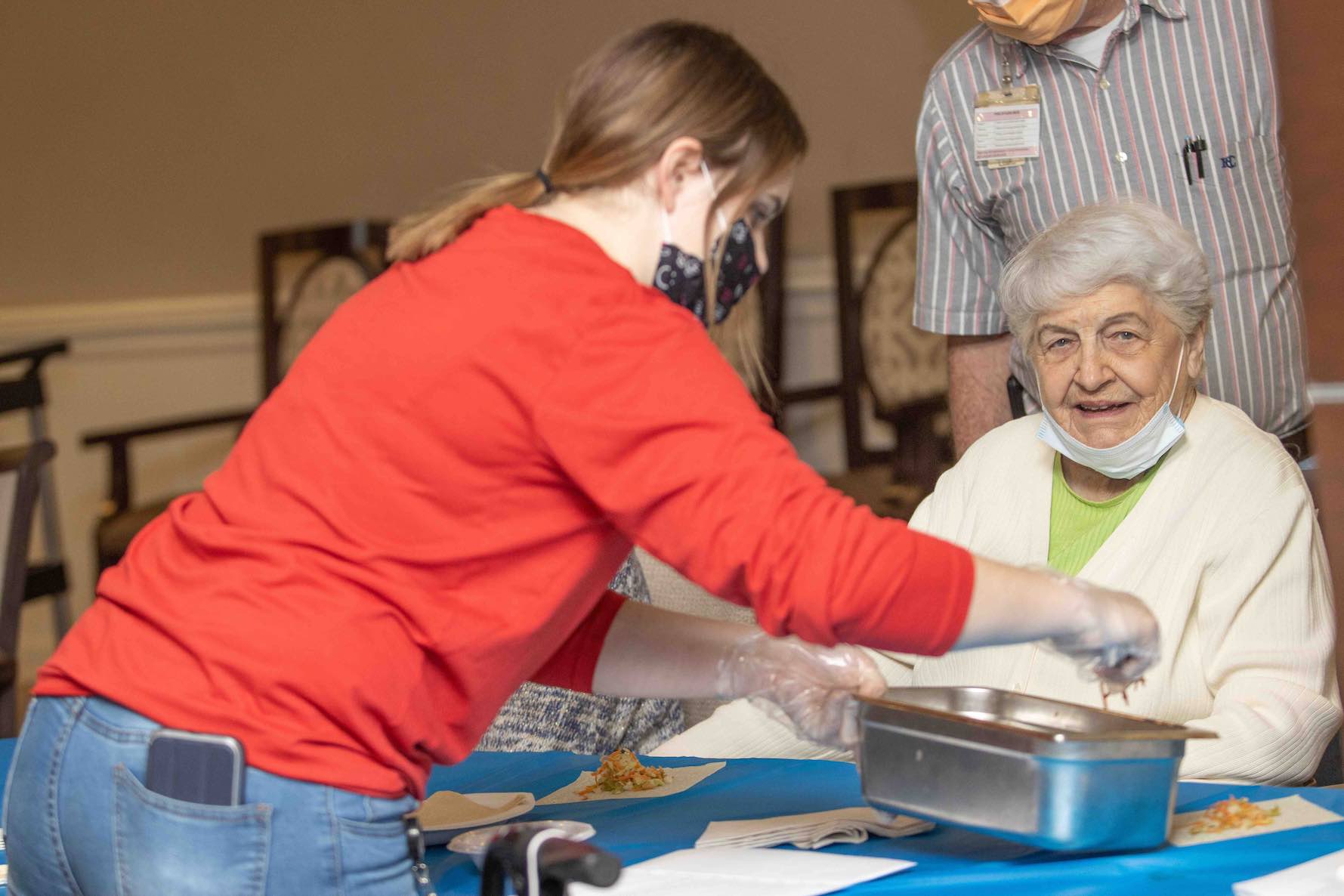 A woman in a red sweatshirt working with another woman to prep food on a blue tablecloth.