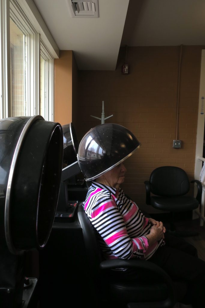 An older woman sitting with her head inside a dome hair dryer.