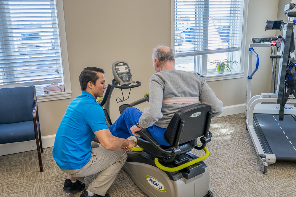 A physical therapist kneels beside an elderly man on a stationary bicycle.