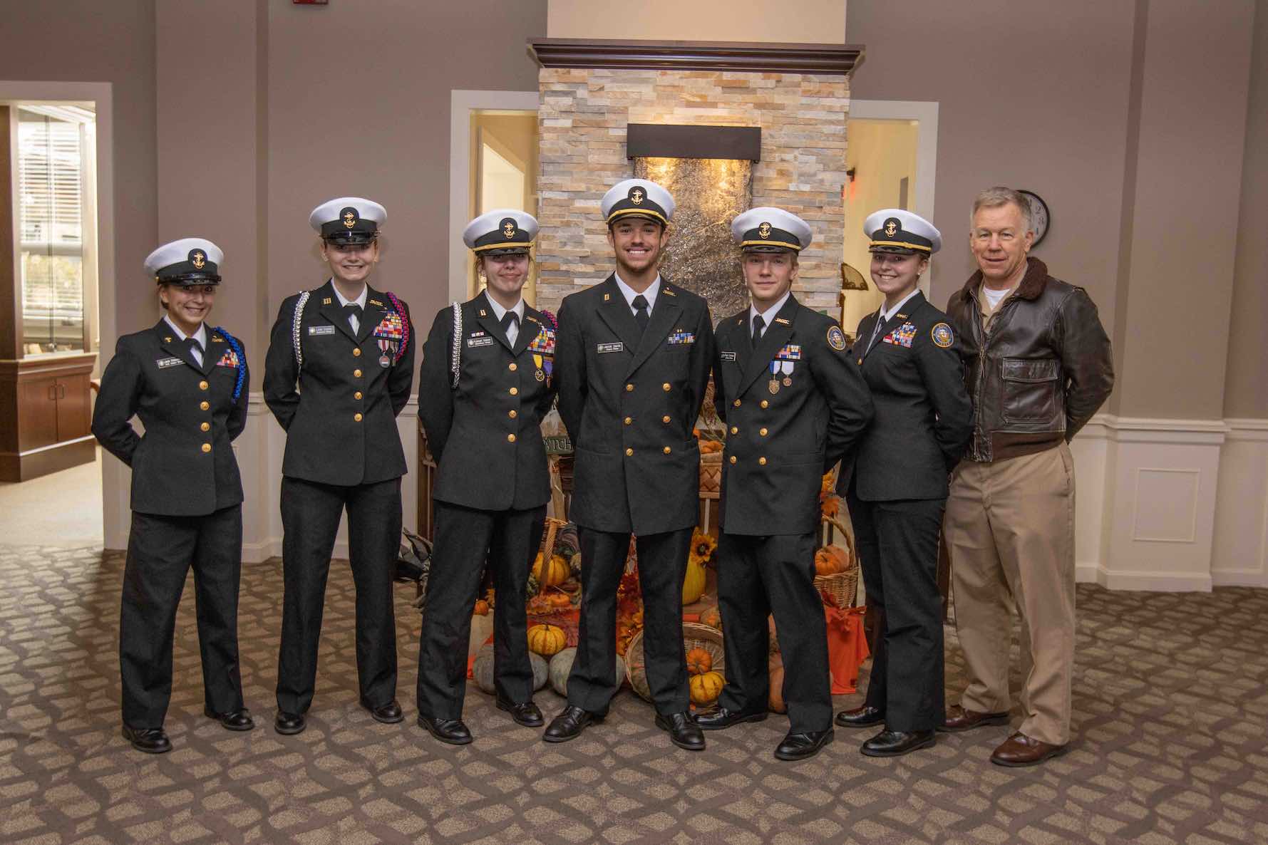 Six Navy Junior ROTC members in uniform with an older adult in a leather bomber jacket.