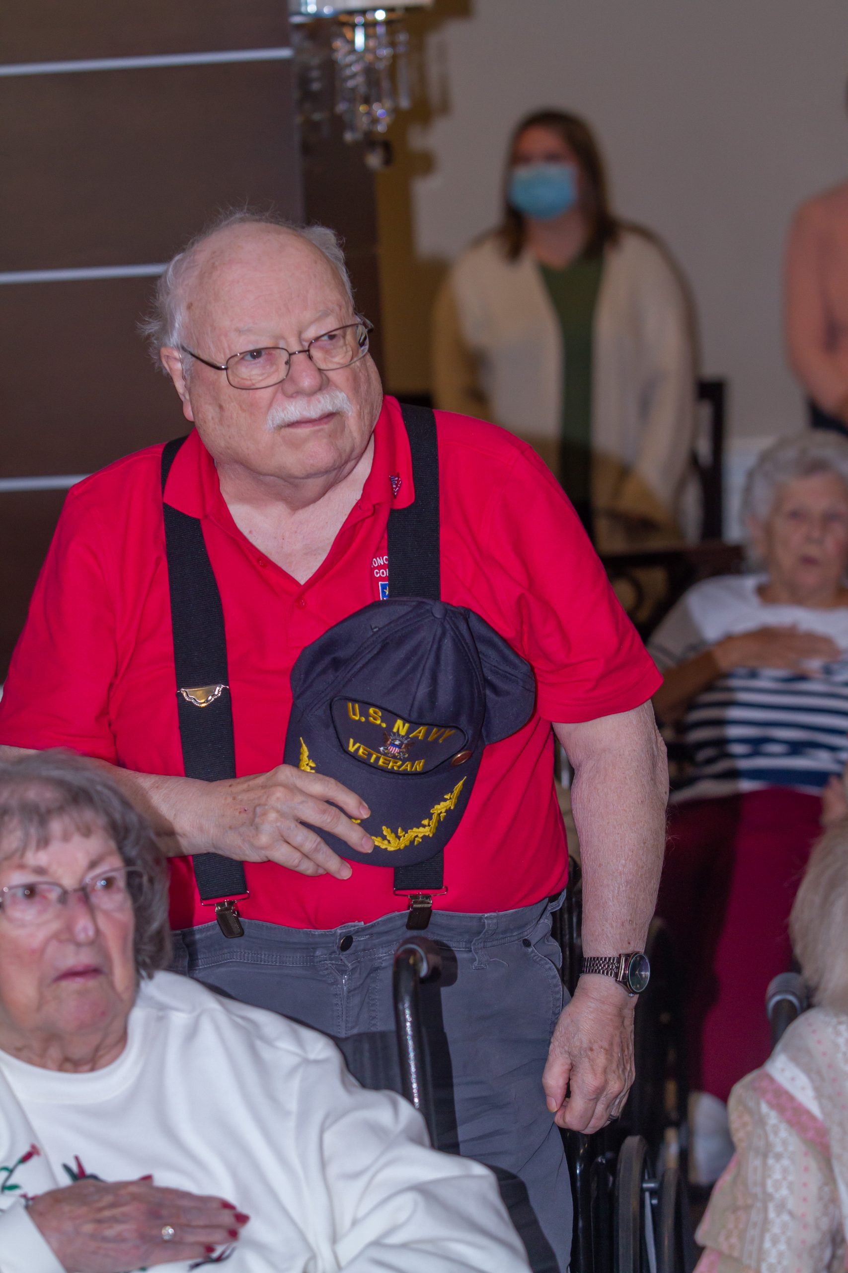 A veteran in overalls holds his hat in his right hand against his heart.