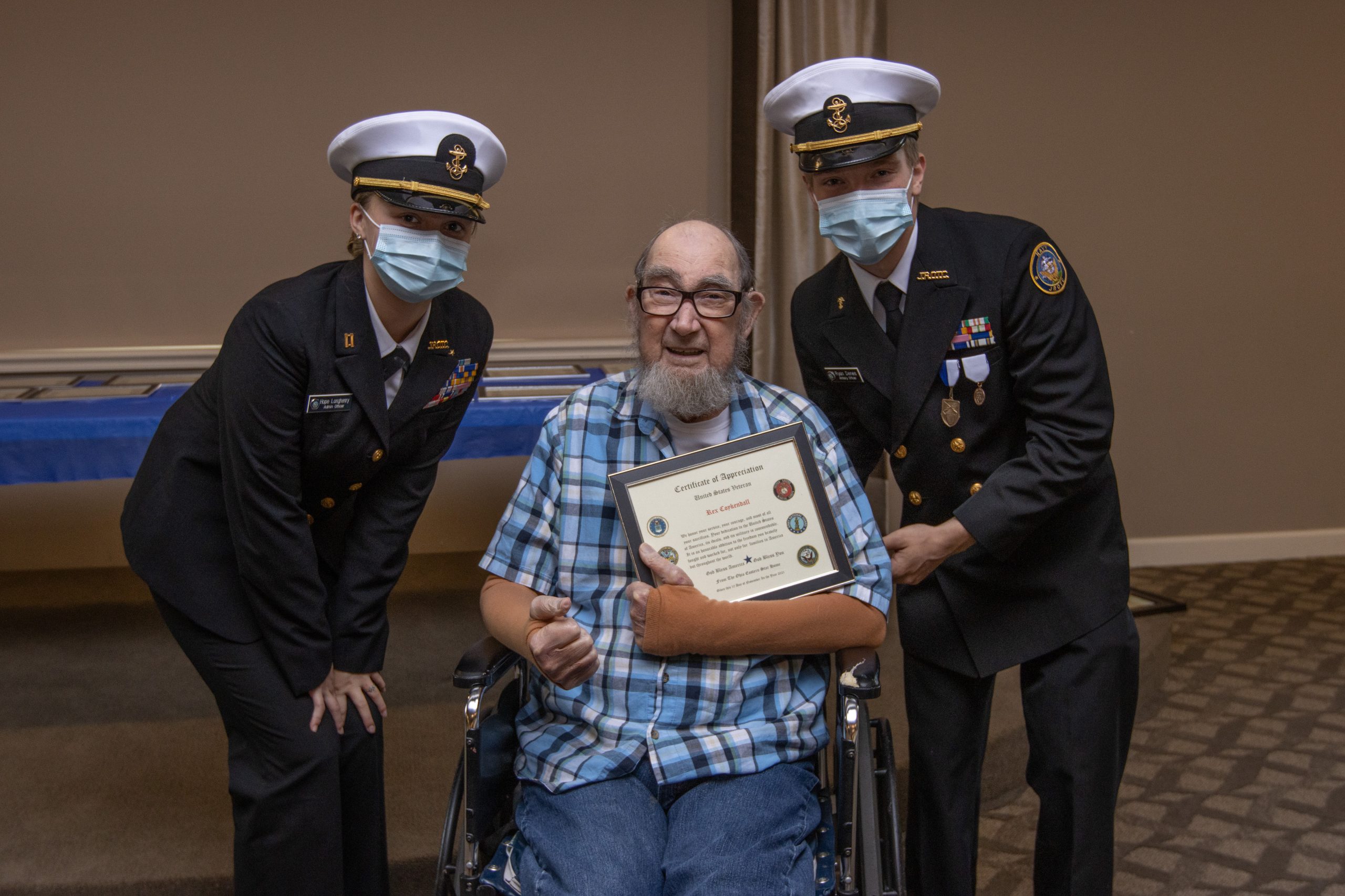 Two uniformed Navy Junior ROTC members in PPE masks flank a senior veteran holding a certificate and sitting in a wheelchair.