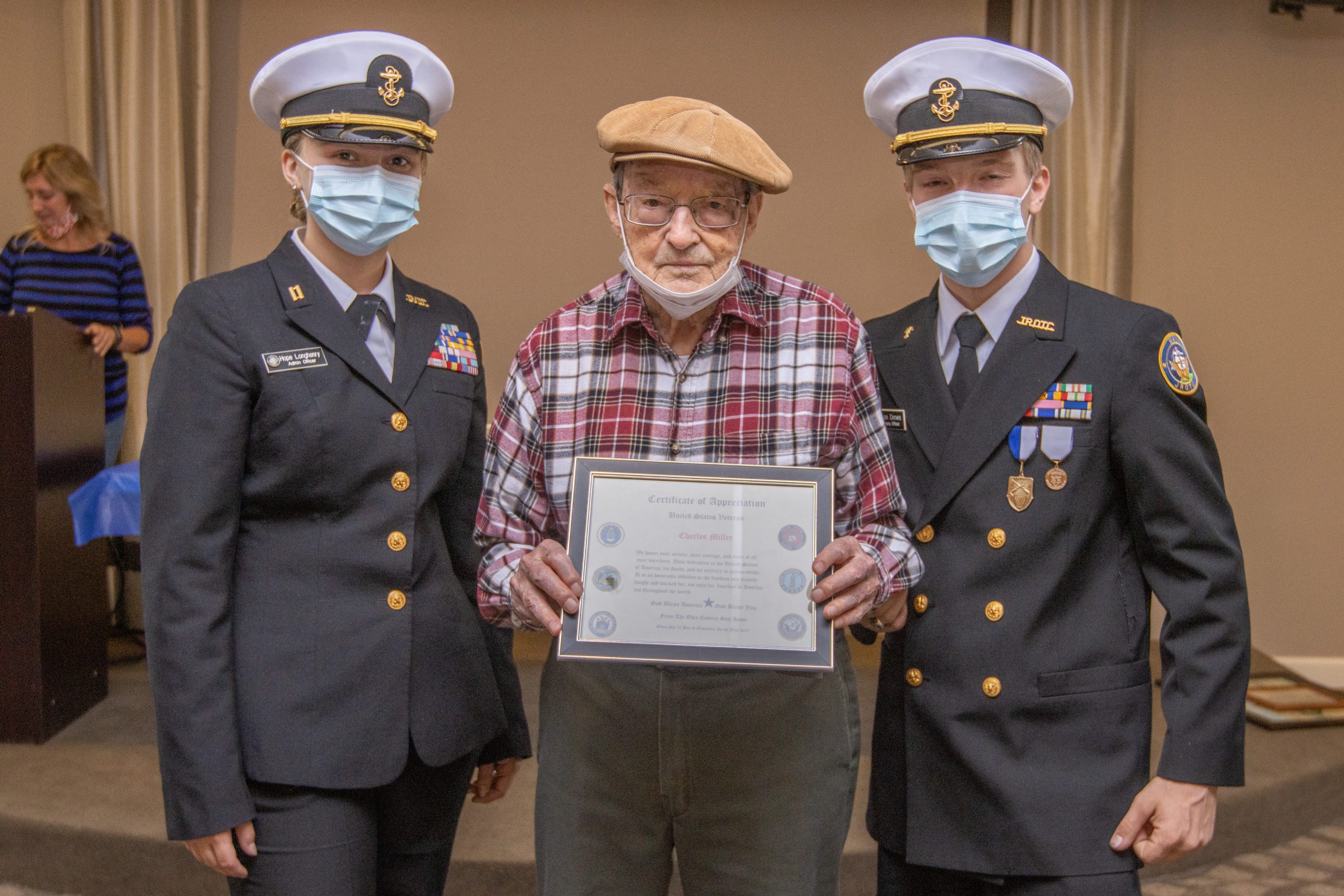 A veteran in a tan beret holds a certificate between two Navy Junior ROTC members.