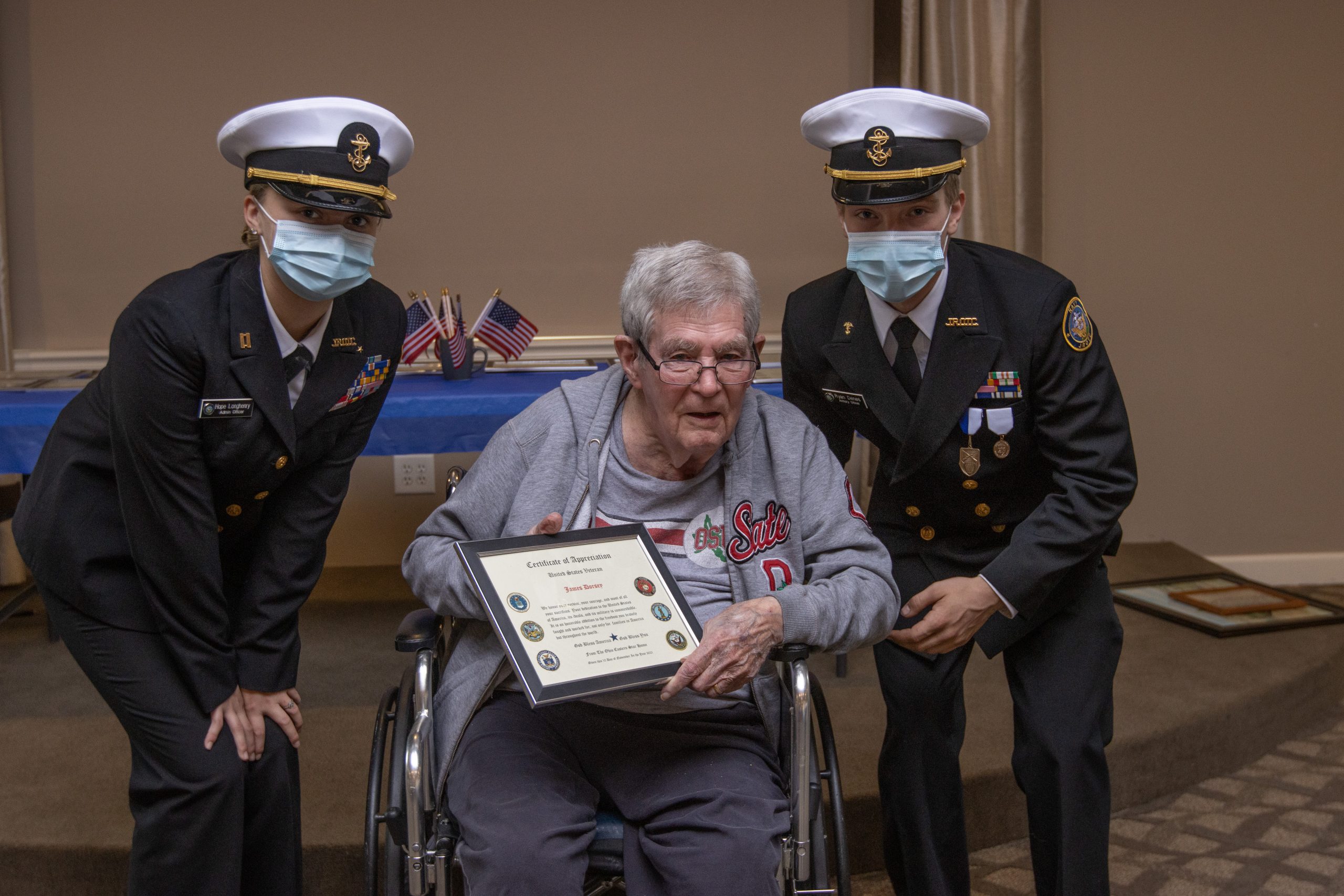 Two uniformed Navy Junior ROTC members in PPE masks flank a grey-haired veteran holding a certificate and sitting in a wheelchair.