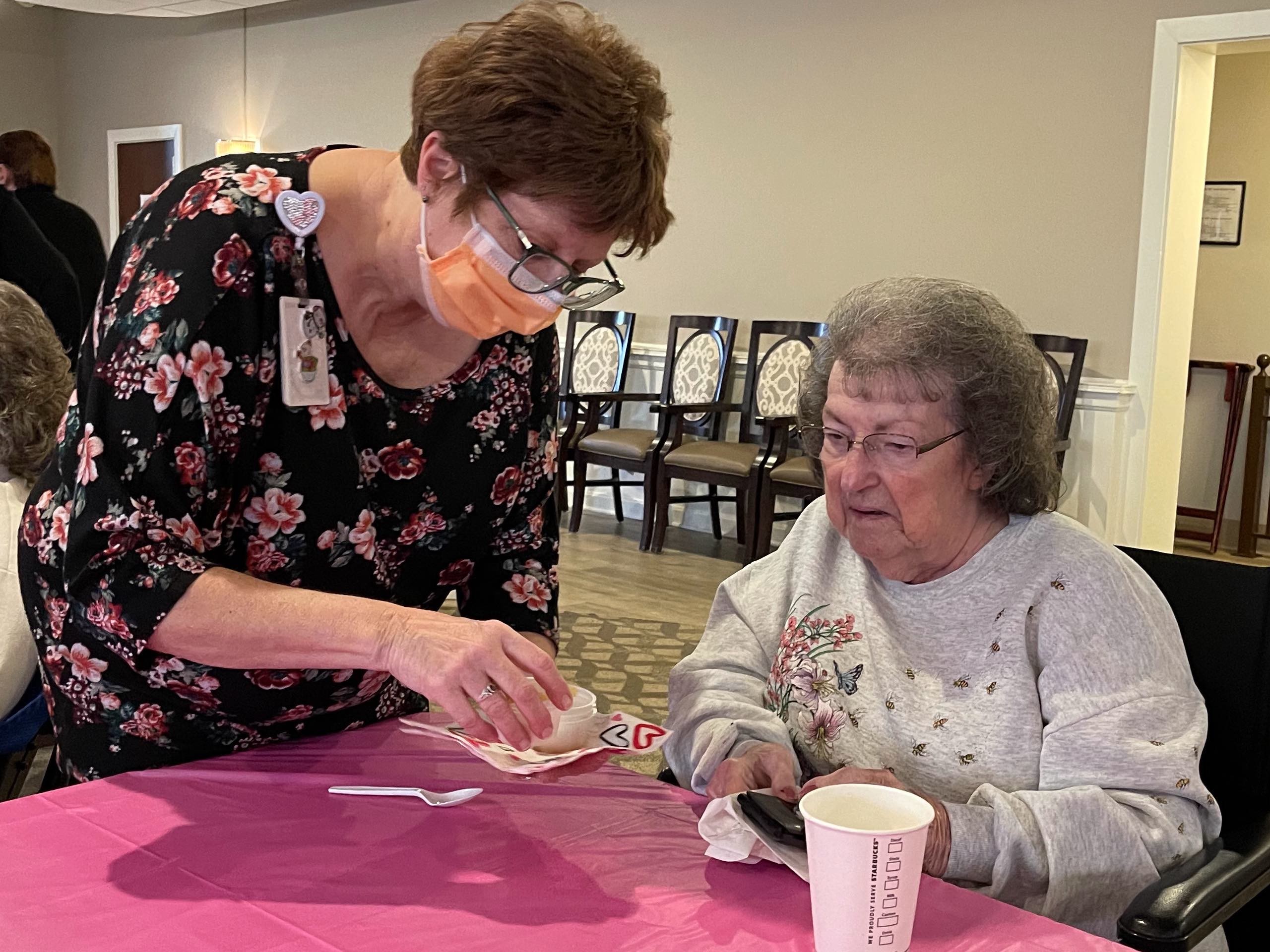 A woman in a floral shirt and mask sets some soup in front of a seated elder.