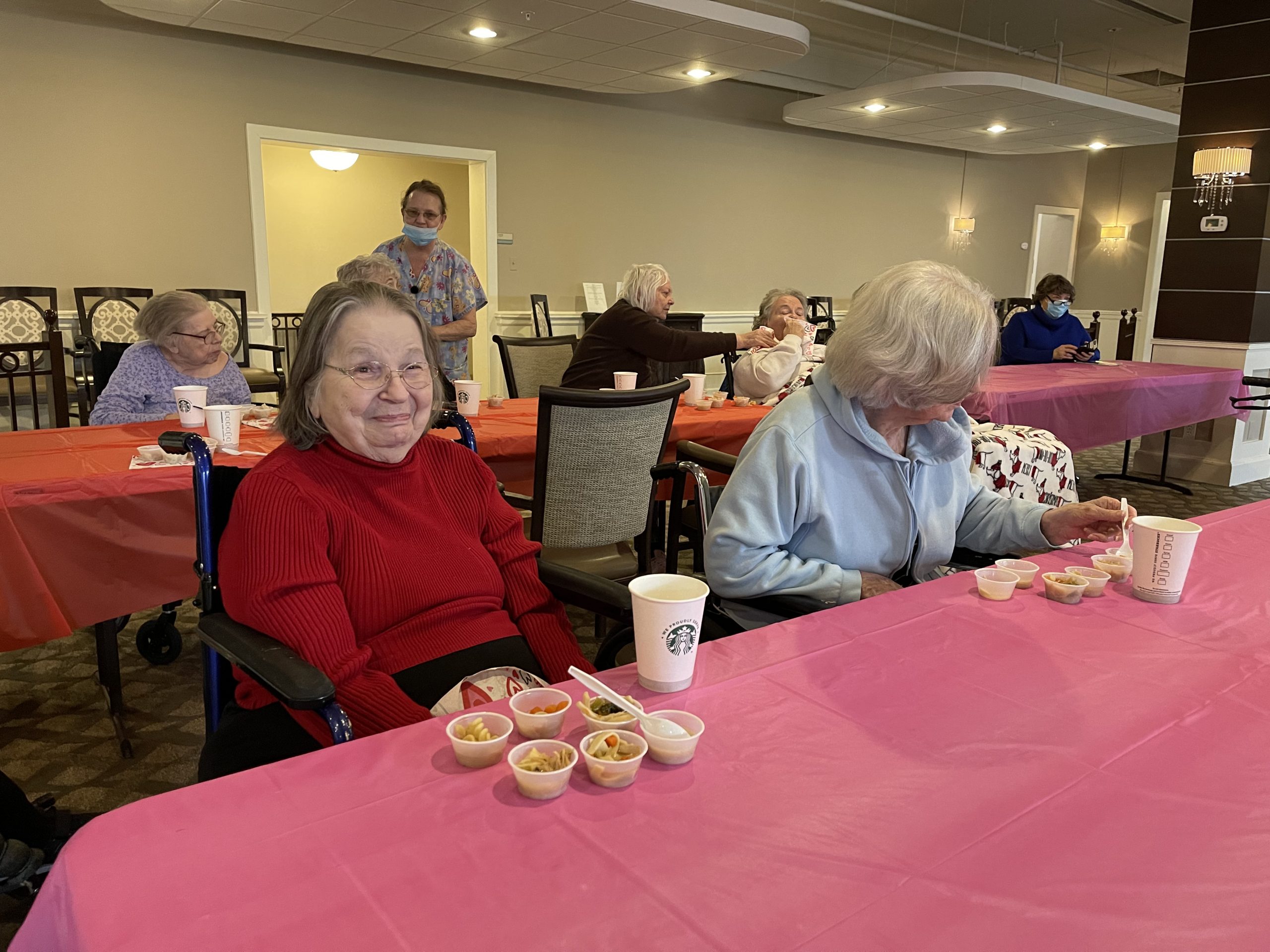 A woman in a red sweater smiles at the camera in a room of elderly folks eating soup.