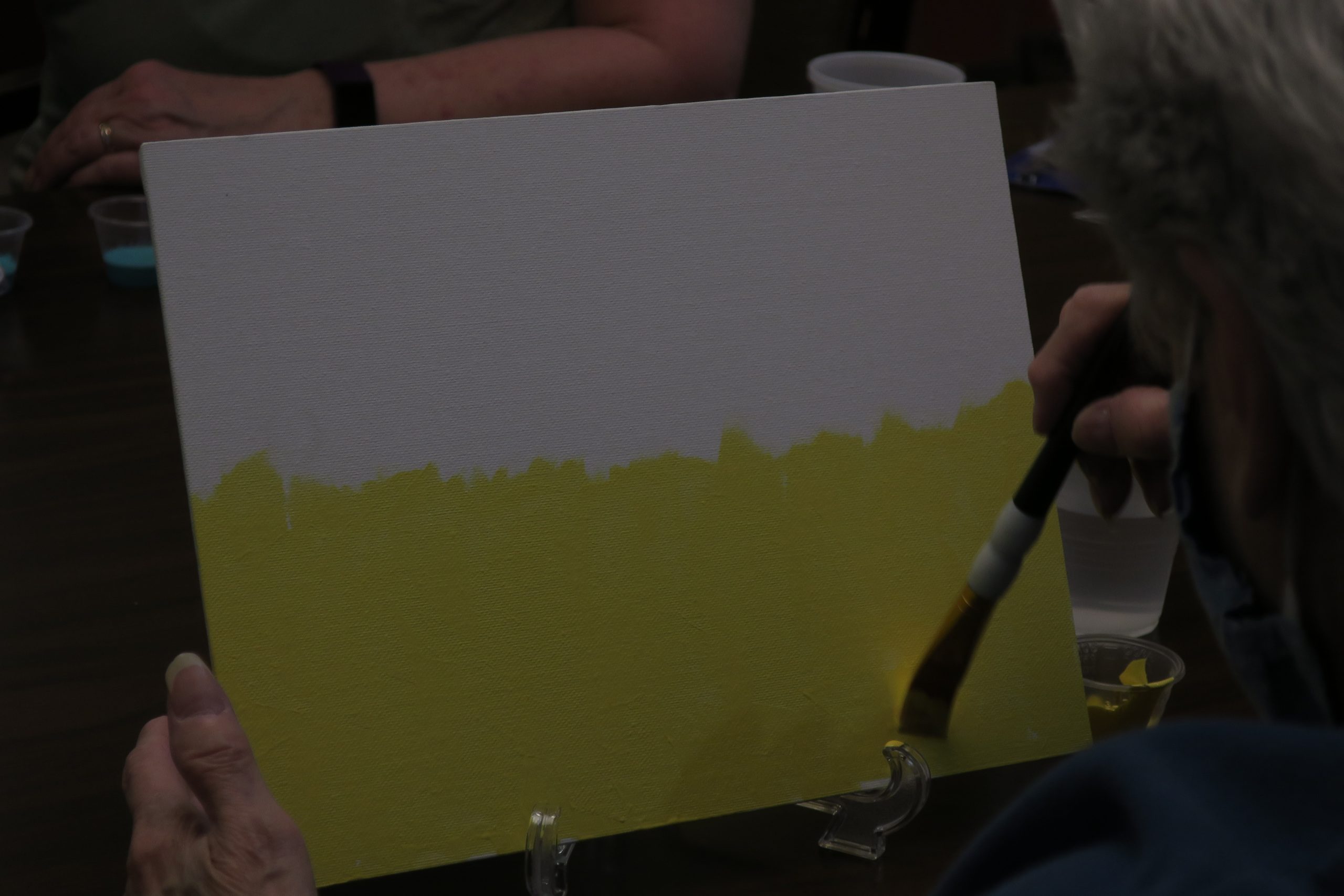 An elderly person paints yellow on a white canvas.
