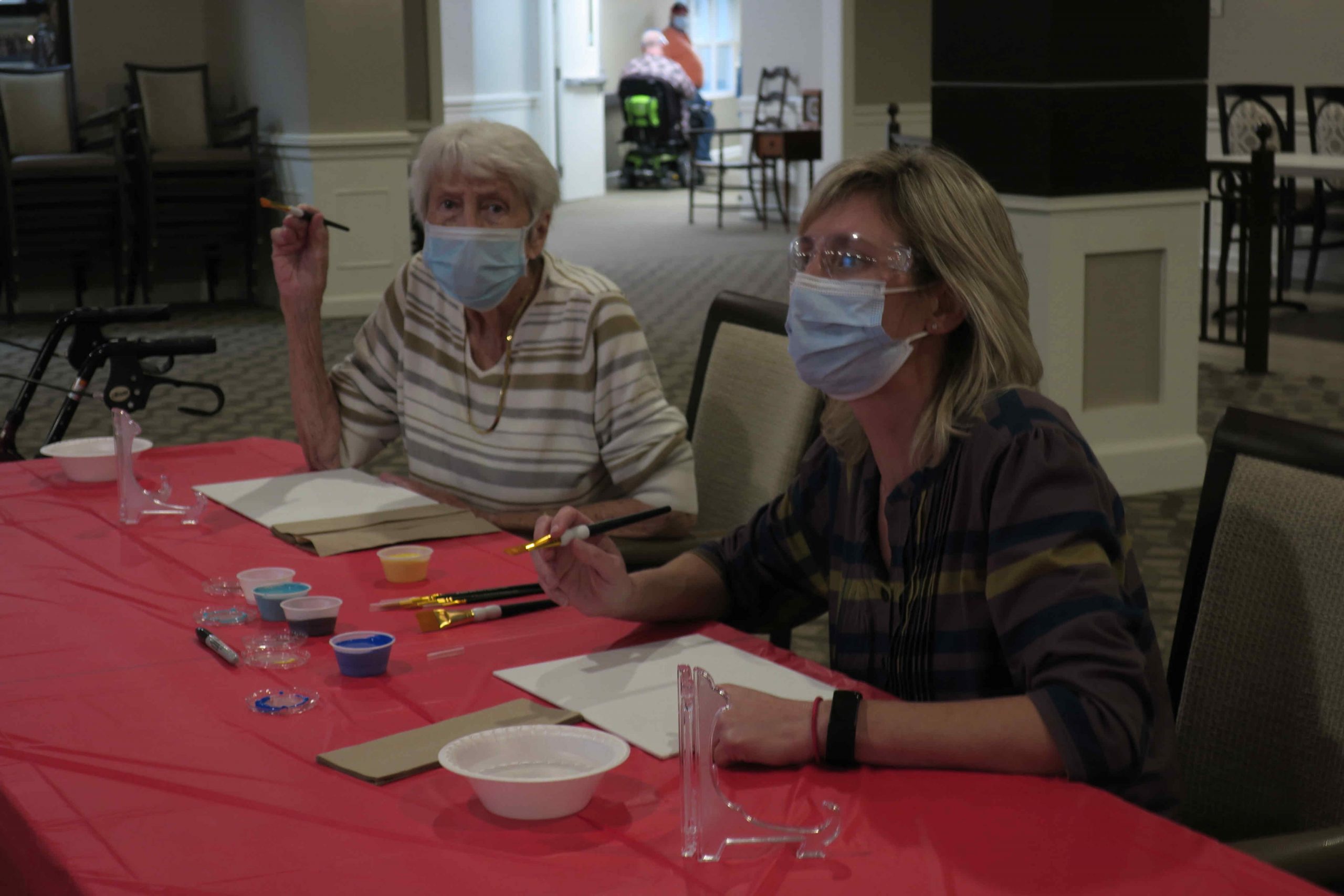 Two women painting on a red arts and crafts table with masks on.