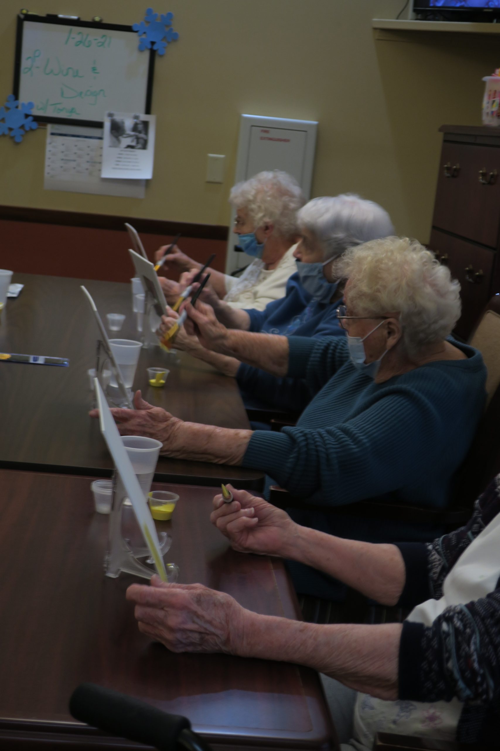 Four elderly women sitting at a dark, wooden table are painting.