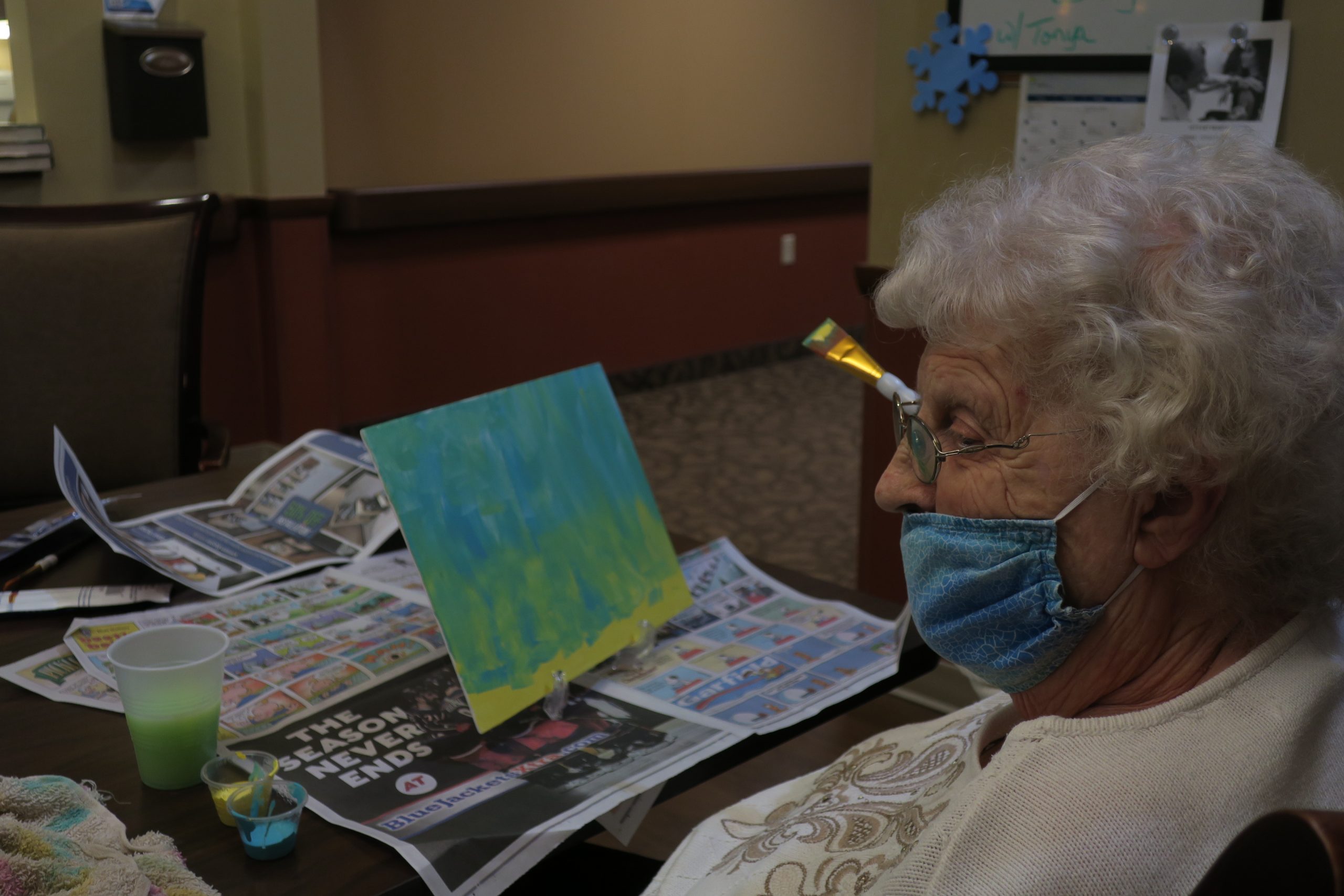A woman with white, wavy hair sits at a table in front of a blue, green, and yellow painting.