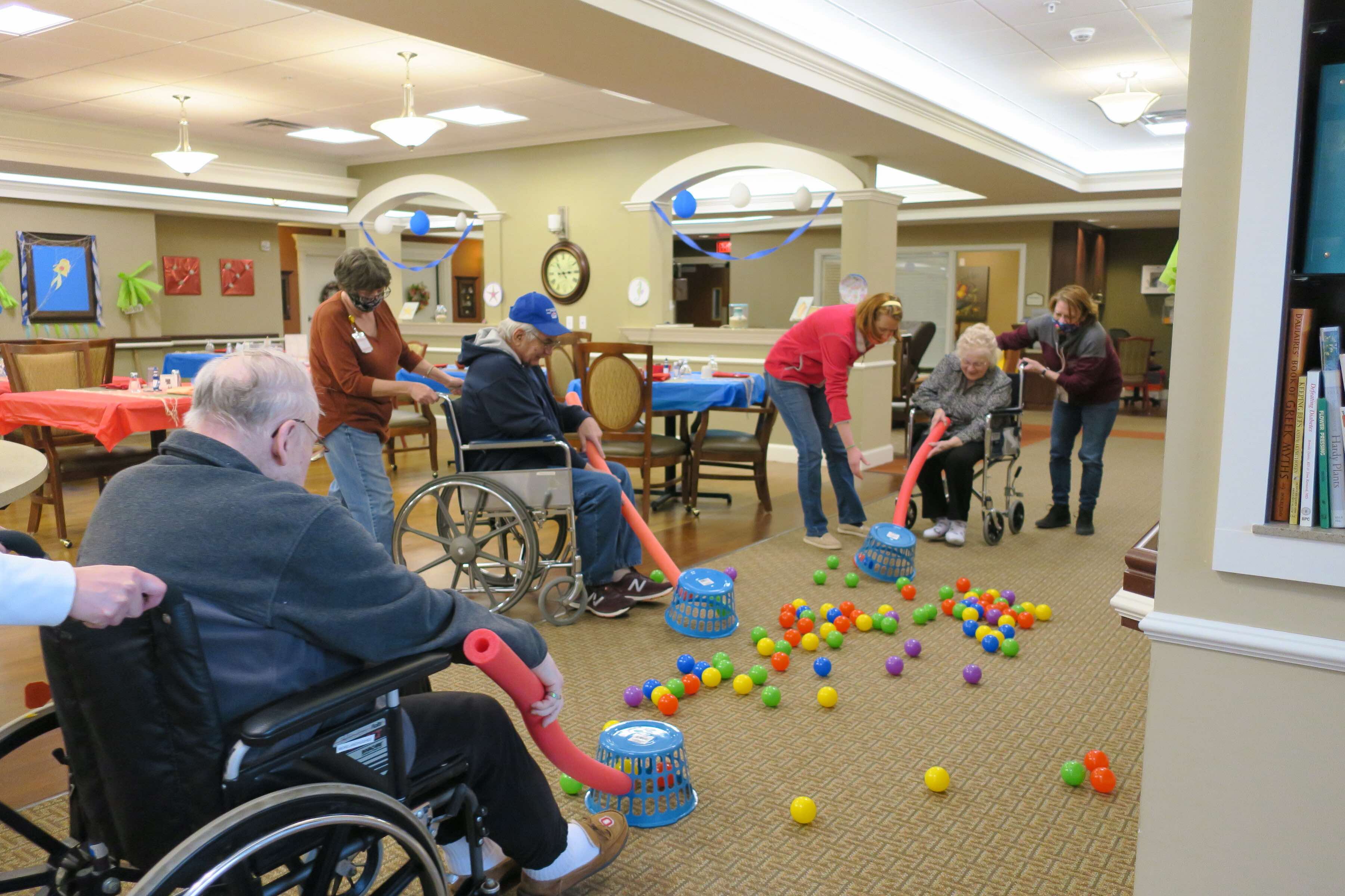 Nursing home residents play a fun and silly game with pool noodles, plastic balls, and laundry baskets while stuck indoors during COVID-19.