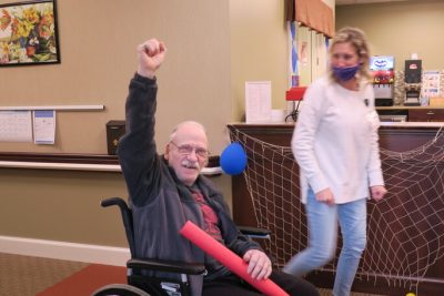 An older man in a wheelchair raises a fist and holds a red pool noodle in his other hand.