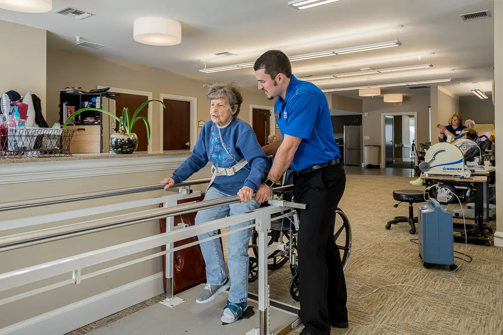 An occupational therapy professional supports an elderly lady on parallel bars equipment.