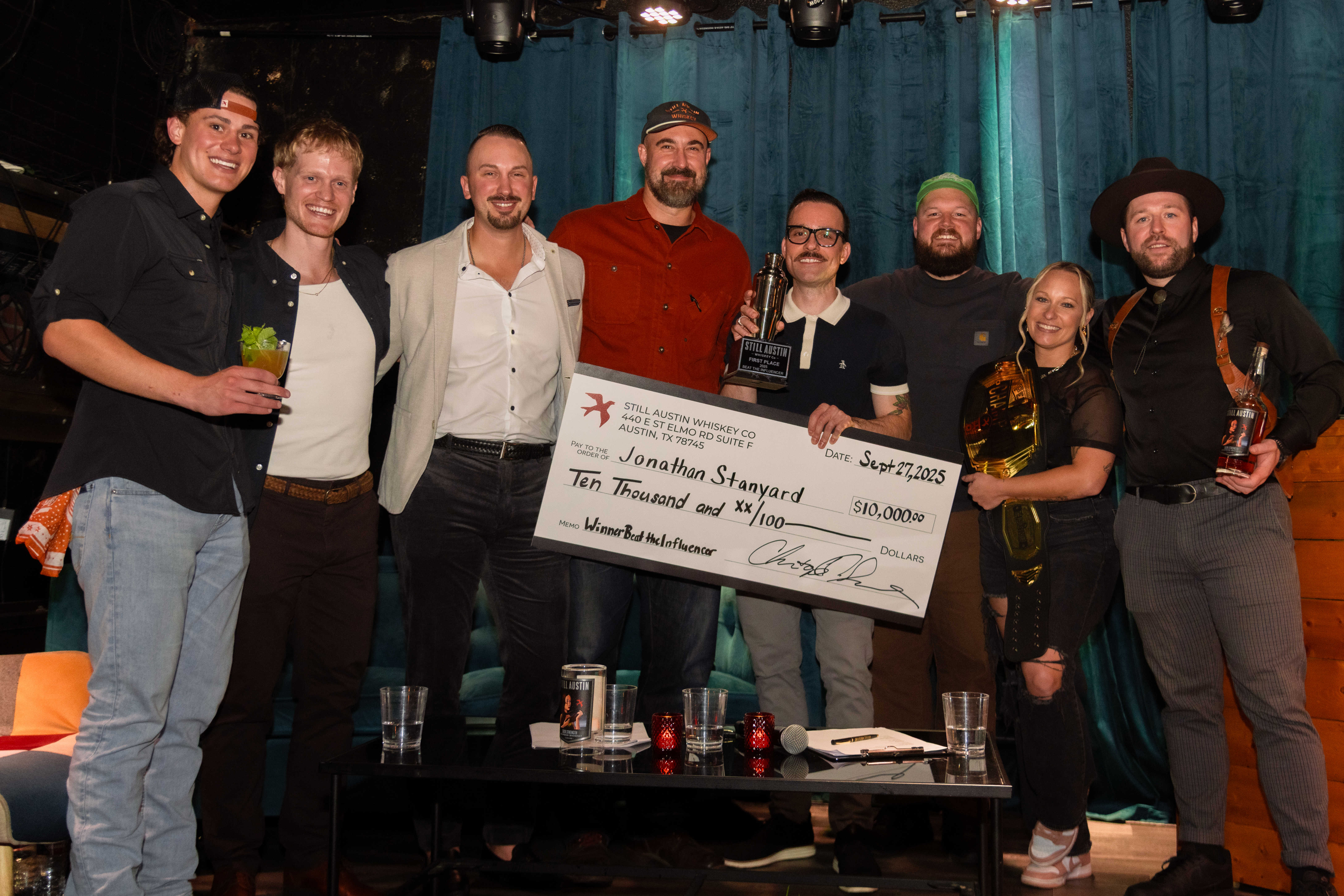 Beat The Influencer winners and judges celebrating on stage — Jonathan with the big check, Ashley rocking the championship belt, and the whole crew raising a glass.