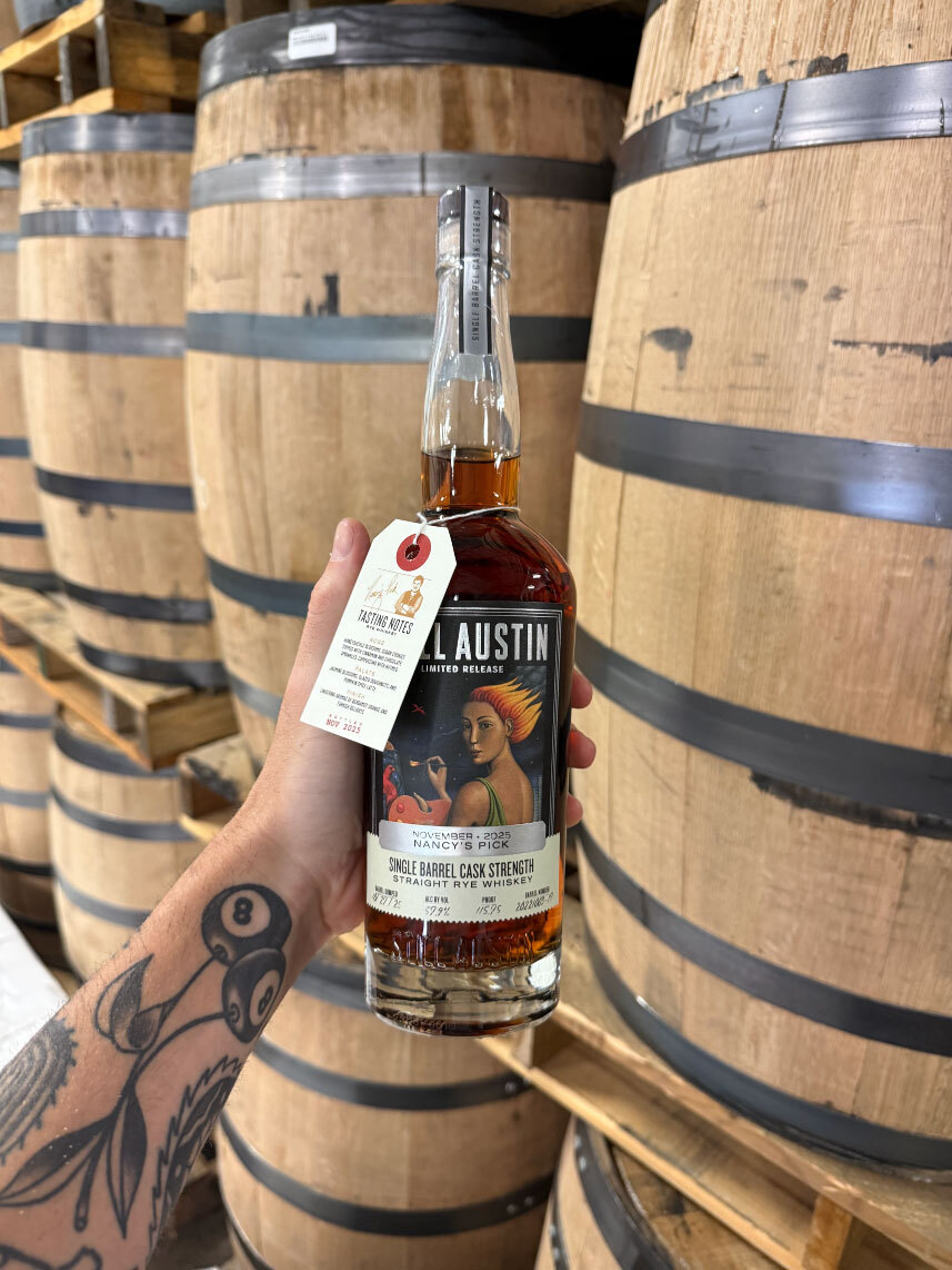 A tattooed hand holds a bottle of Still Austin Whiskey Co.’s November 2025 Nancy’s Pick Single Barrel Cask Strength Straight Rye Whiskey in front of stacked oak barrels inside the Austin distillery.