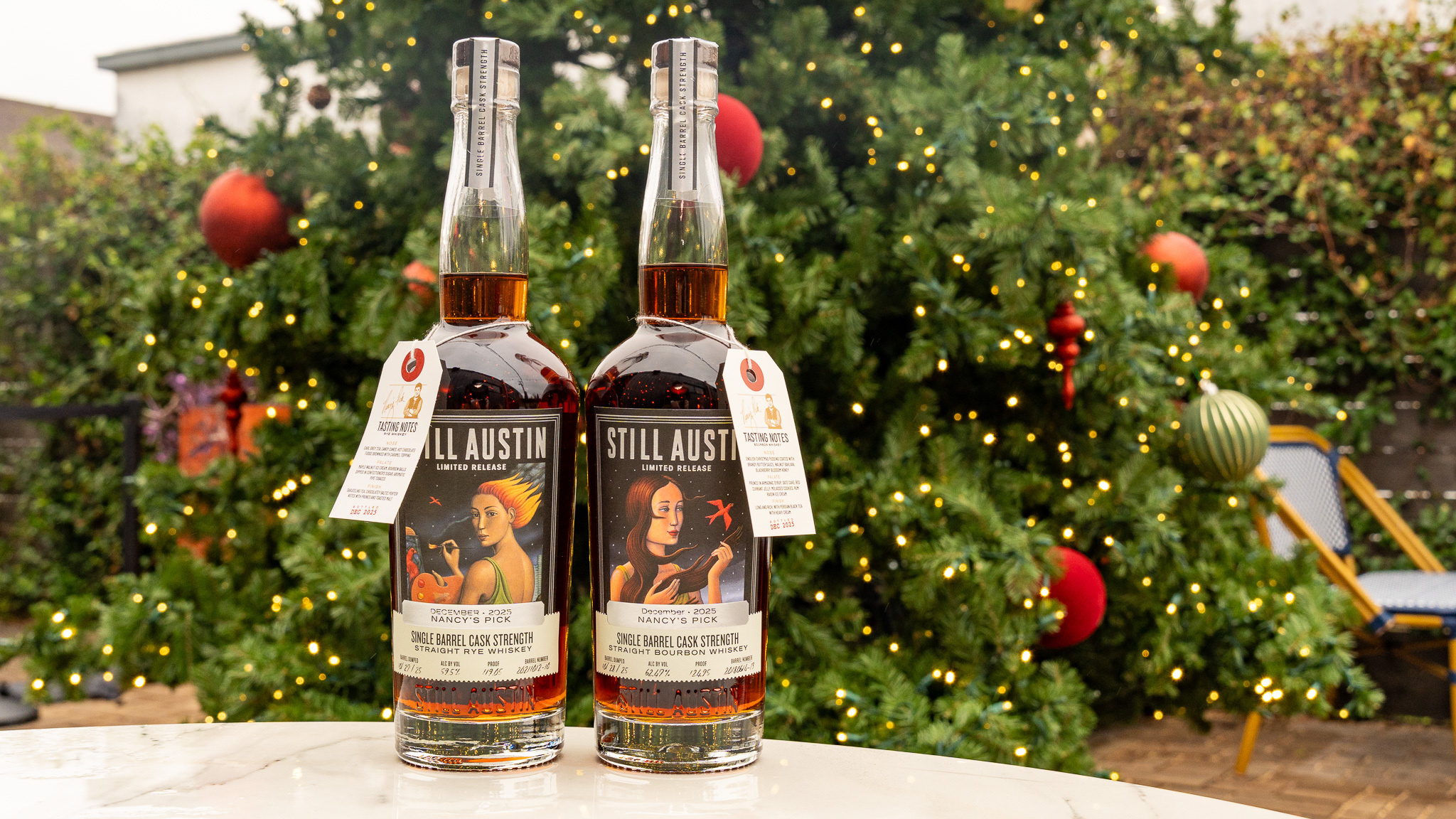 Two limited-release Still Austin Nancy’s Picks bottles —one rye and one bourbon— stand on a white table in front of a large Christmas tree decorated with warm lights and colorful ornaments in an outdoor setting.