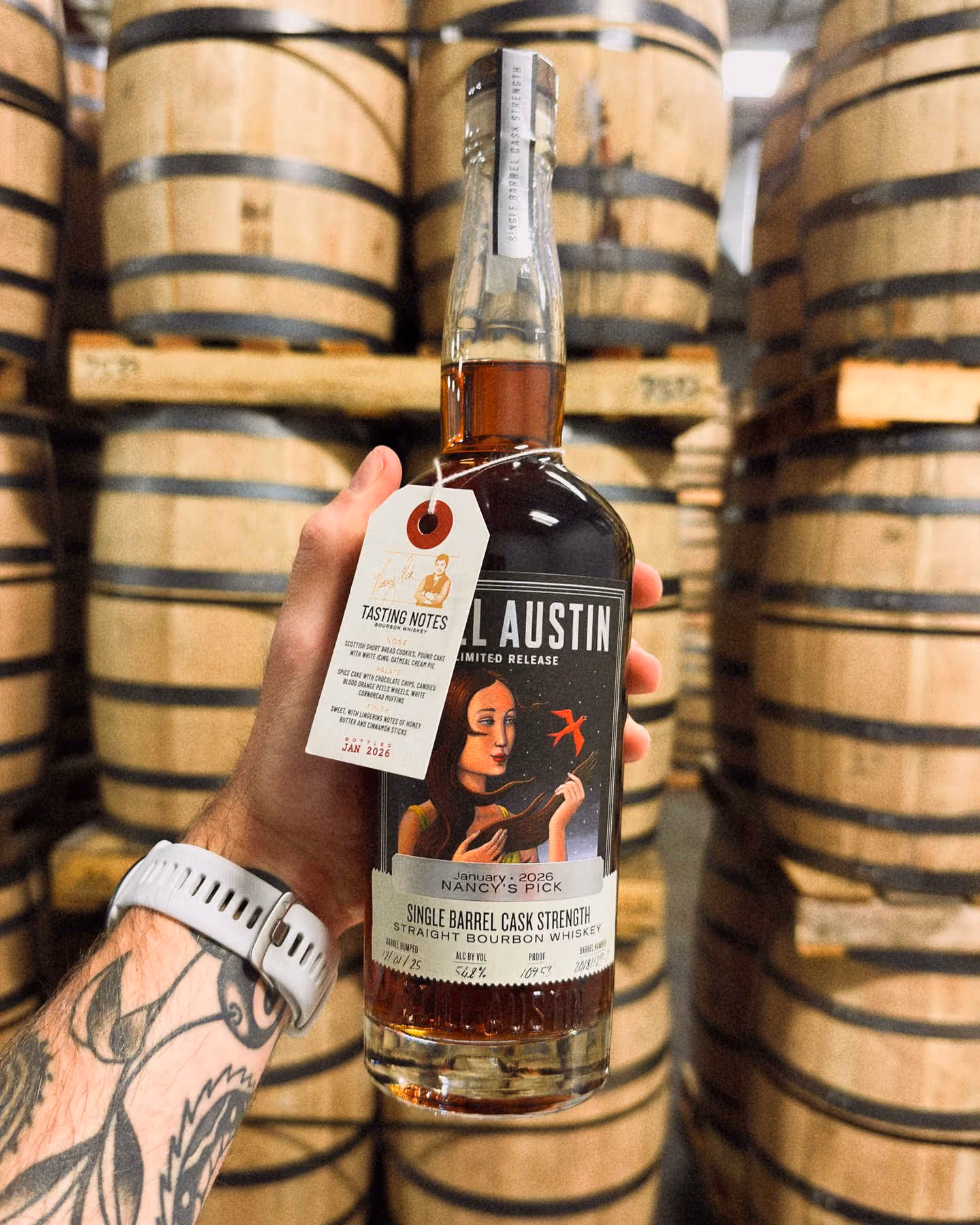 Hand holding a bottle of Still Austin January 2026 Nancy’s Pick Single Barrel Cask Strength Straight Bourbon Whiskey in front of stacked aging barrels inside the rickhouse.