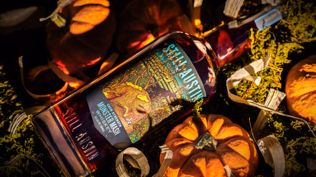bottle of Still Austin Monster Mash Whiskey with a Halloween-themed label featuring a mummy with pumpkins. The bottle has a wooden cork and a black ribbon.