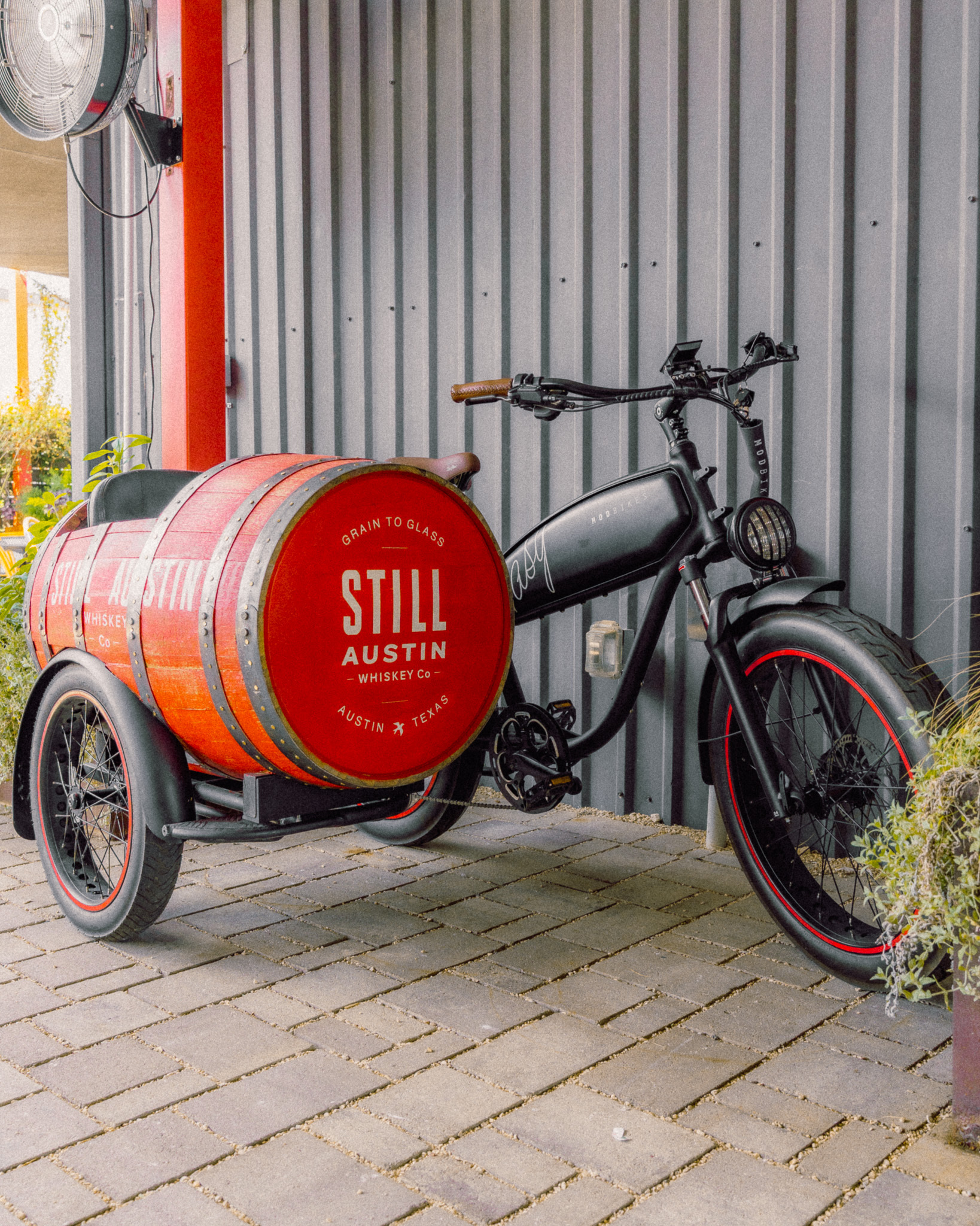 A custom Still Austin Whiskey Co. cargo e-bike with a red whiskey barrel sidecar is parked outside the distillery.
