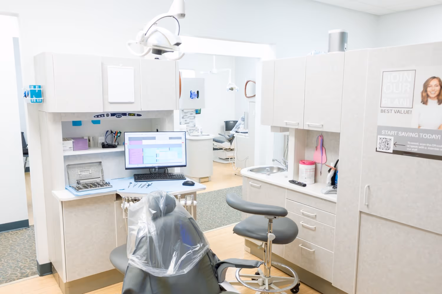 Modern dental exam room with a patient chair covered in plastic, dental tools, a computer monitor, and cabinetry.