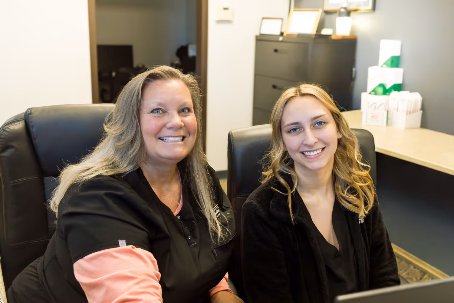 Two women smiling while sitting side by side in an office setting.