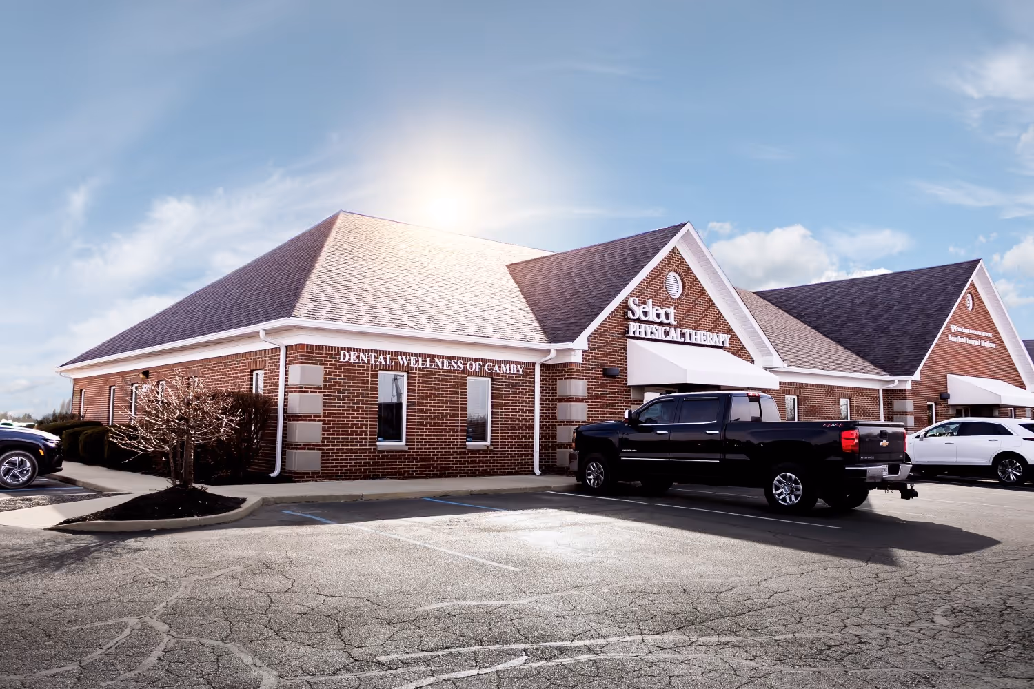 Brick medical office building with signs for Select Physical Therapy and Dental Wellness of Camby, with vehicles parked in front under a bright daytime sky.