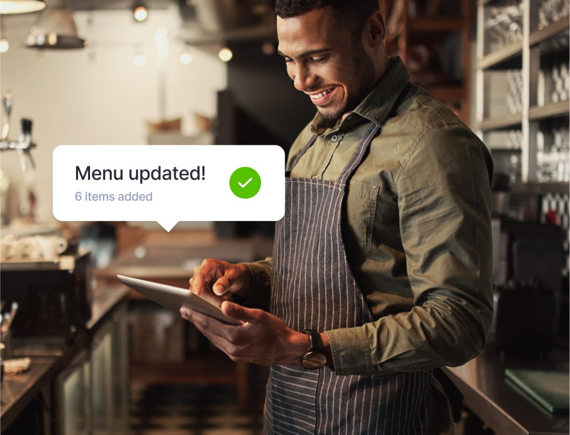Smiling man in an apron updating a menu on a tablet in a kitchen with a notification showing six items added.